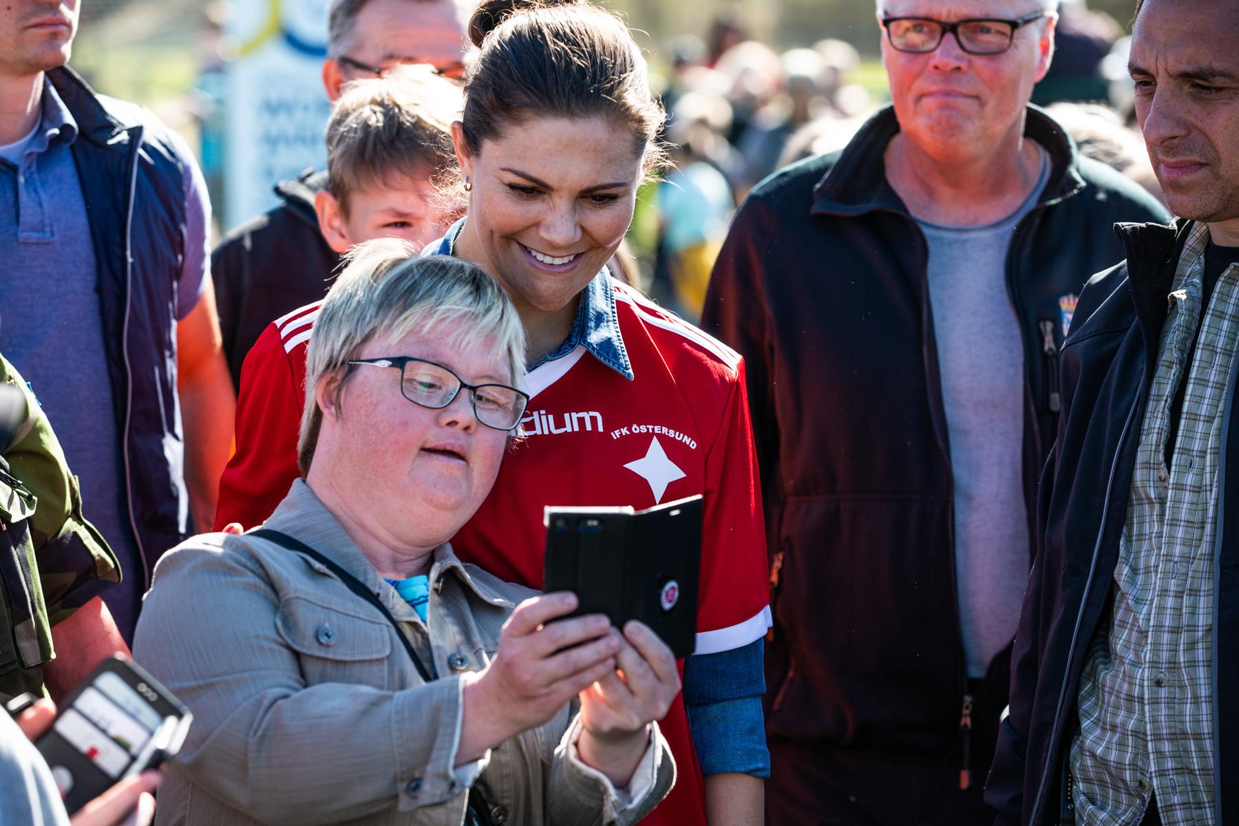 Crown Princess Victoria in a football T-shirt being shown something on a woman's mobile phone. Crown Princess Victoria is a popular member of the Swedish royal family.