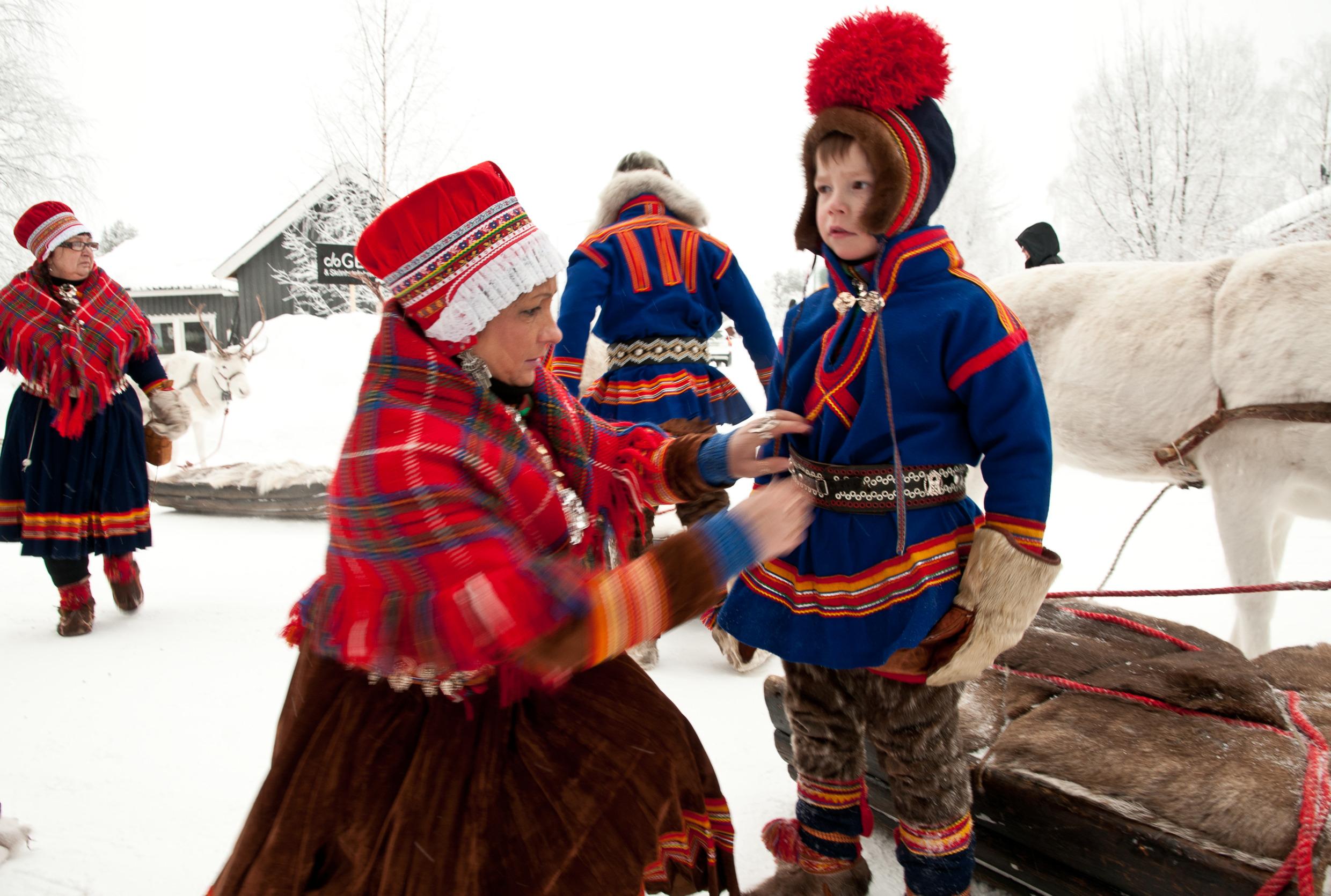 A woman helping a boy dress. They are both wearing traditional Sami clothing. In the background are two more people.