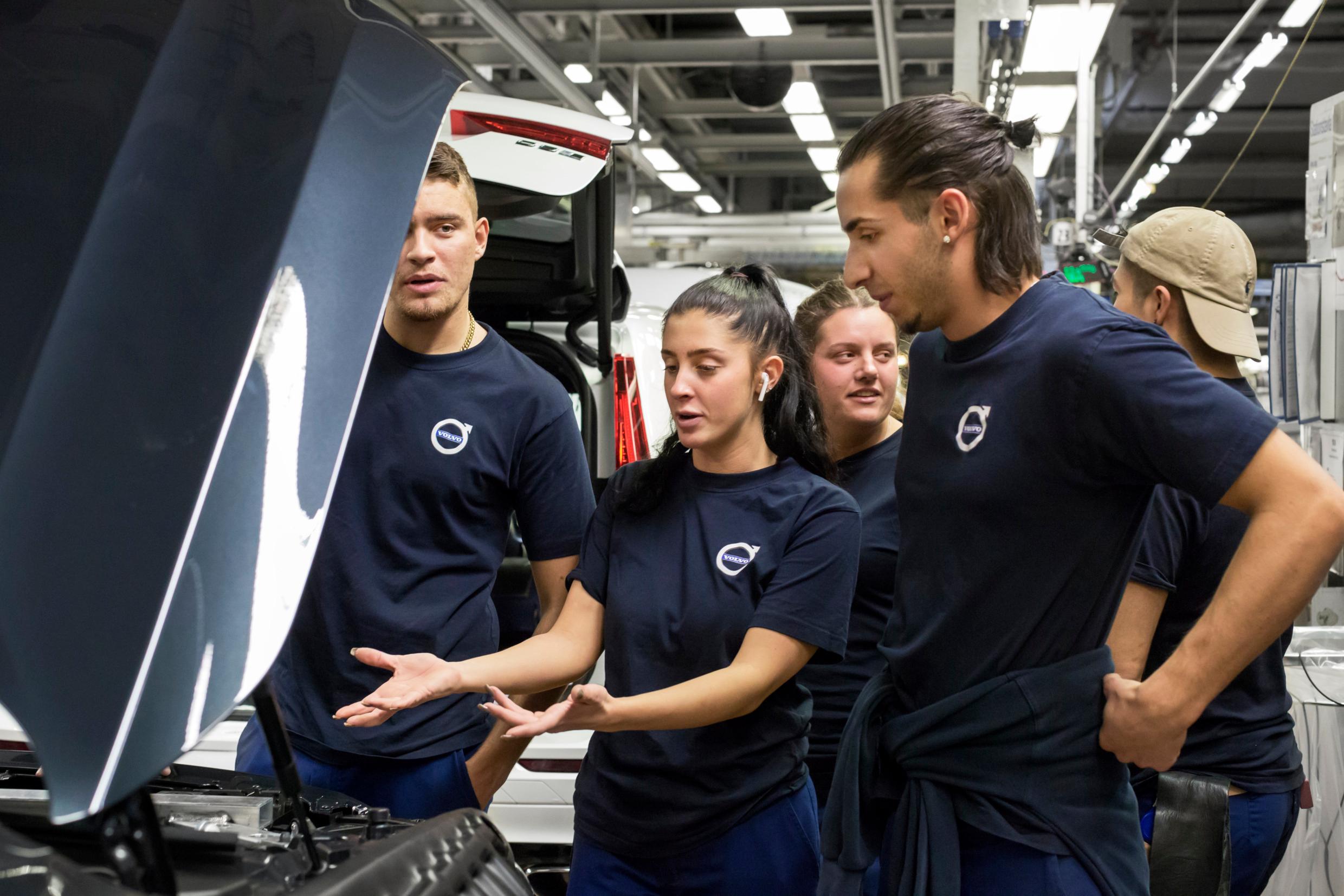 A crowd of workers stand around a car on a factory floor. A woman is gesturing at the engine compartment.