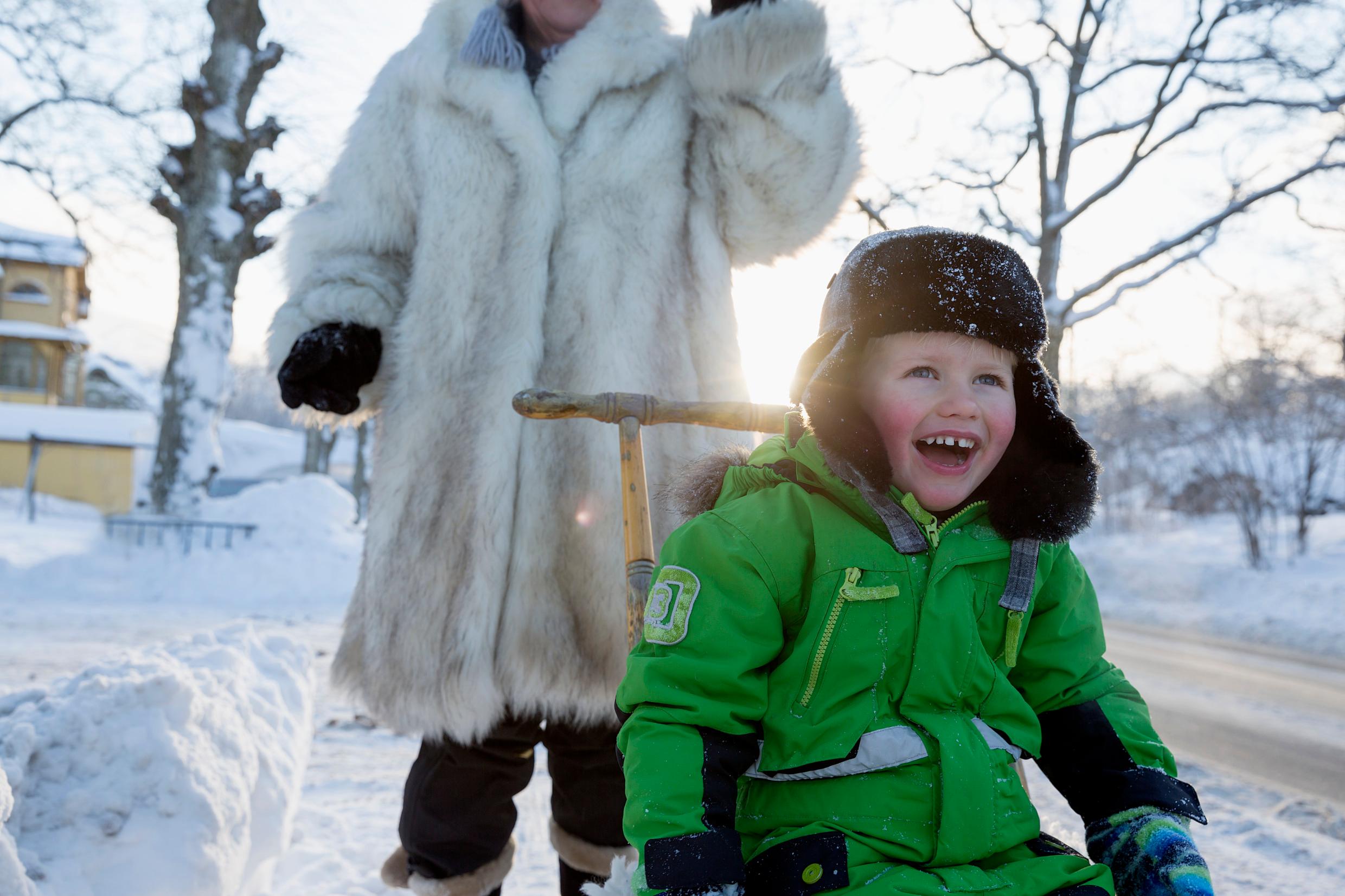 A boy in a green overall and a furry hat on a sledge in the snow. A woman in a fur coat behind him.