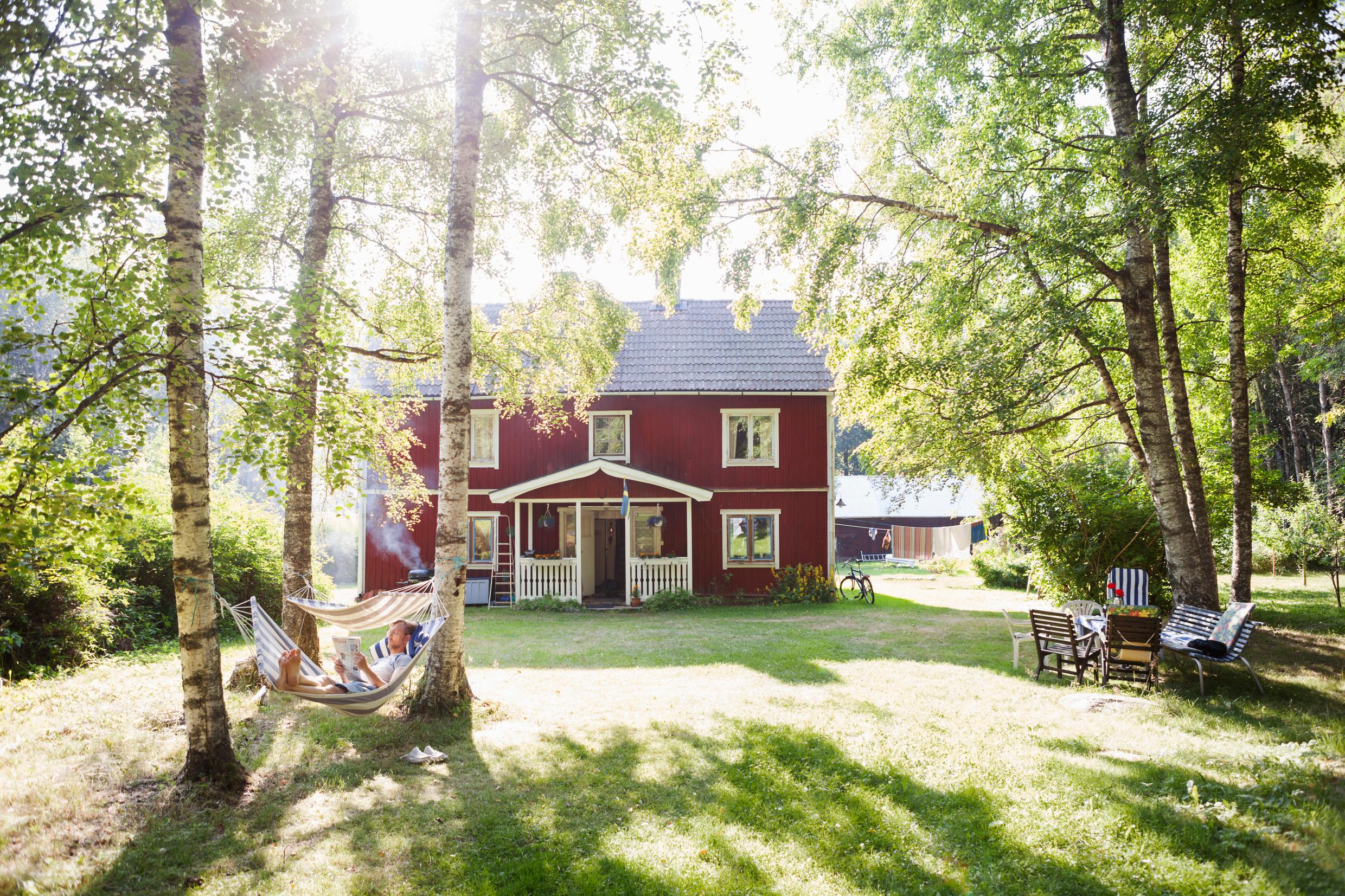 A garden in front of a traditional red Swedish house, with greenery, trees, hammocks and garden furniture.