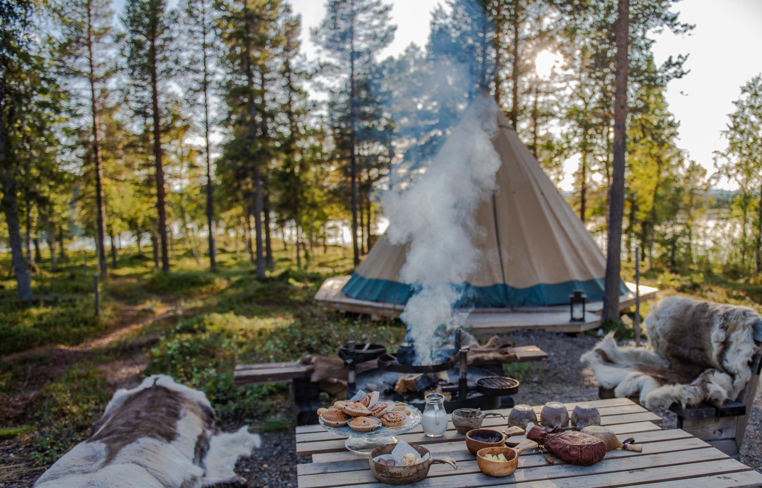 Benches covered in reindeer hides next to a table set with cups and bowls and a plate with buns. A coffee pot is placed over a campfire. A lavvu tent in the background and you get a glimpse of a lake between the trees.