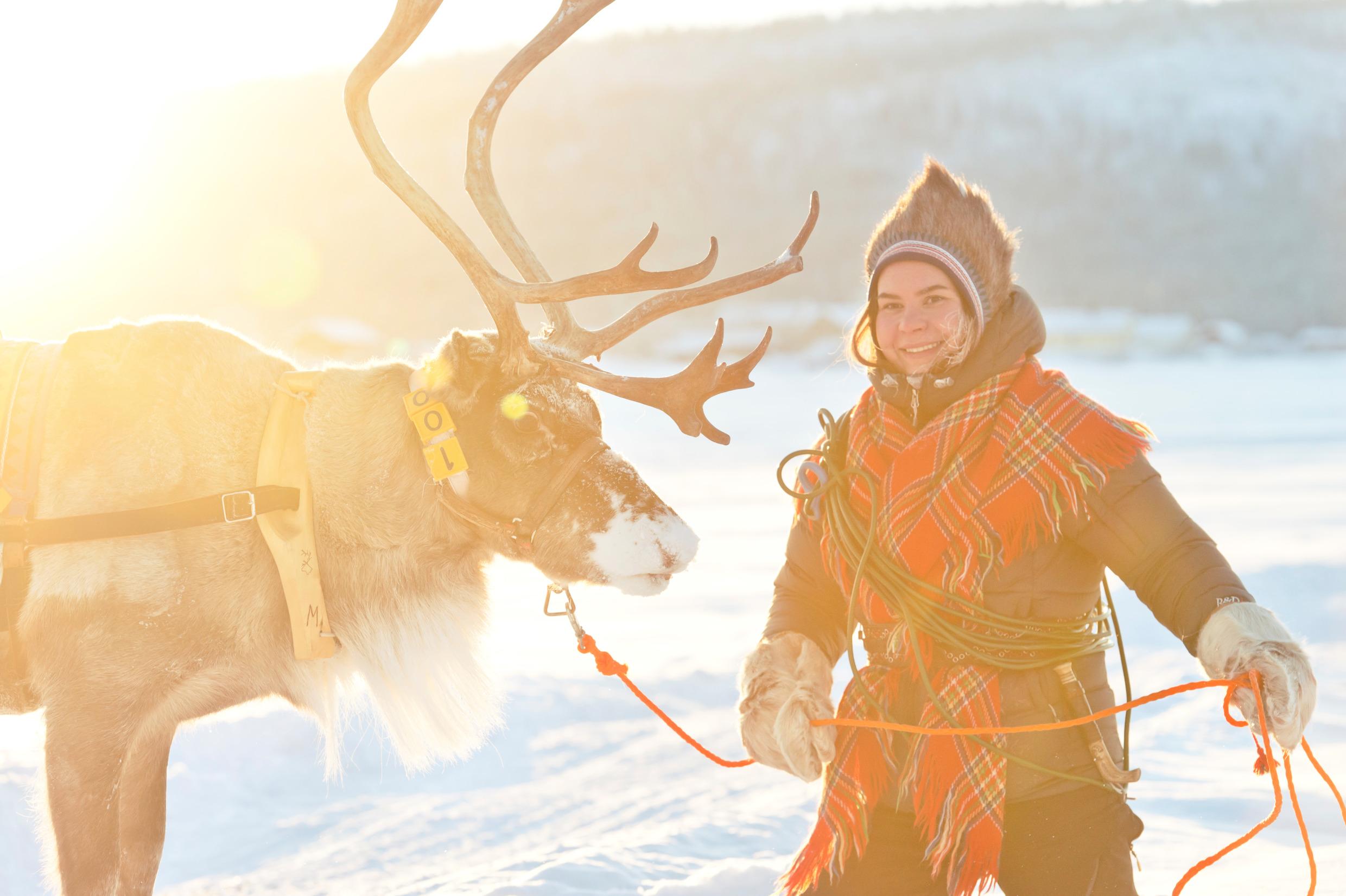 A Sami woman holding a reindeer by a leash, in snowy landscapes.