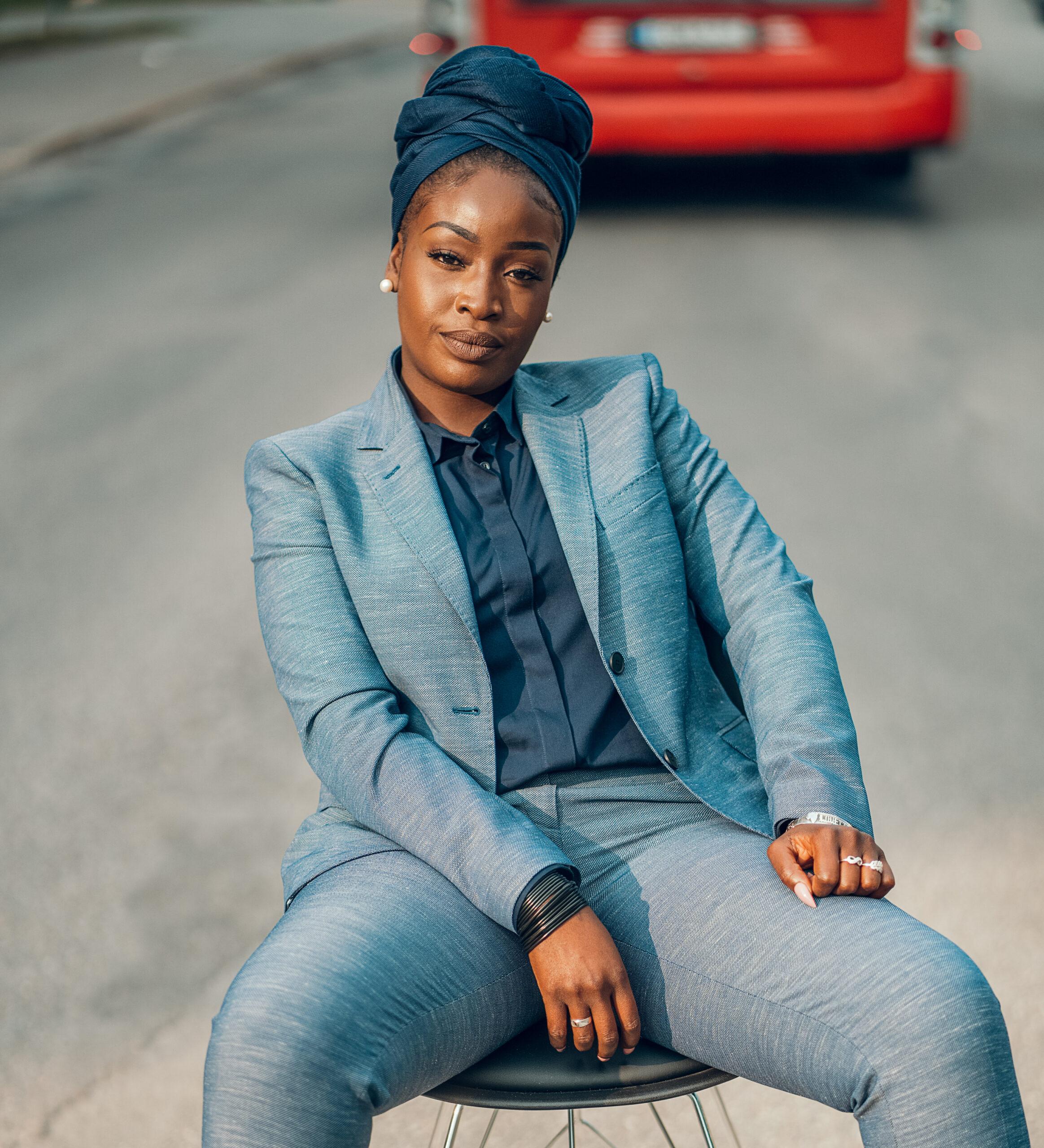 Portrait of Lovette Jallow in a blue suit and a scarf on her head sitting down looking straight into the camera.