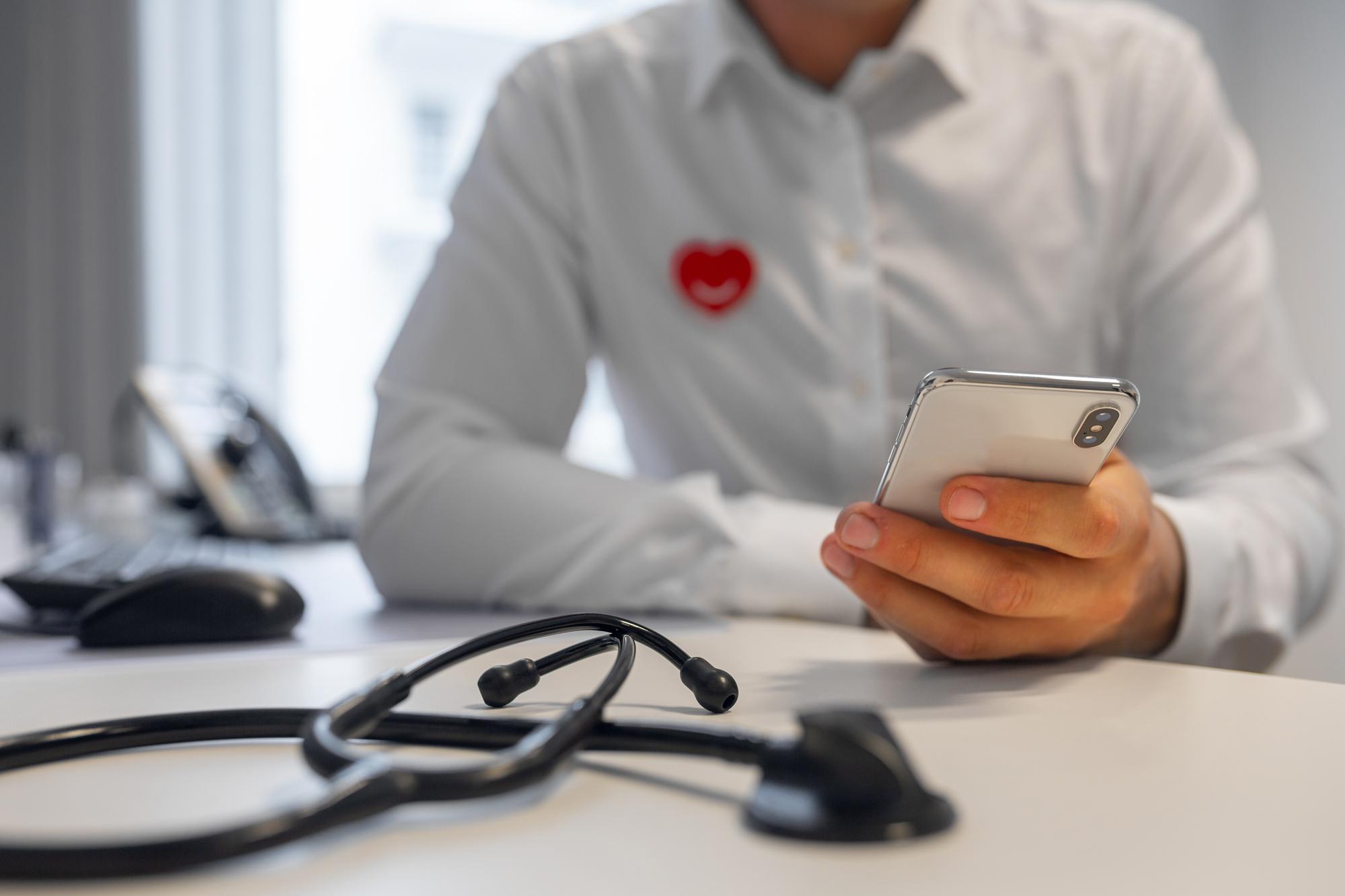 A man in a white shirt seen without his head. He's holding a mobile phone in one hand, on the table in front of him is a stethoscope.