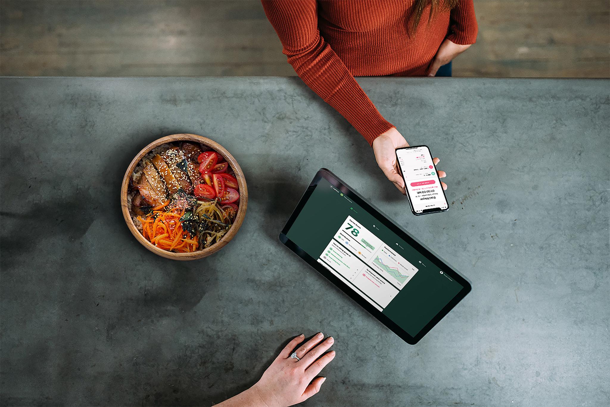 A photo taken from above showing a woman at a counter holding out a hand with a mobile phone. Another person's hand is seen at the bottom of the picture, next to a computer screen. On the counter is also a bowl of food.