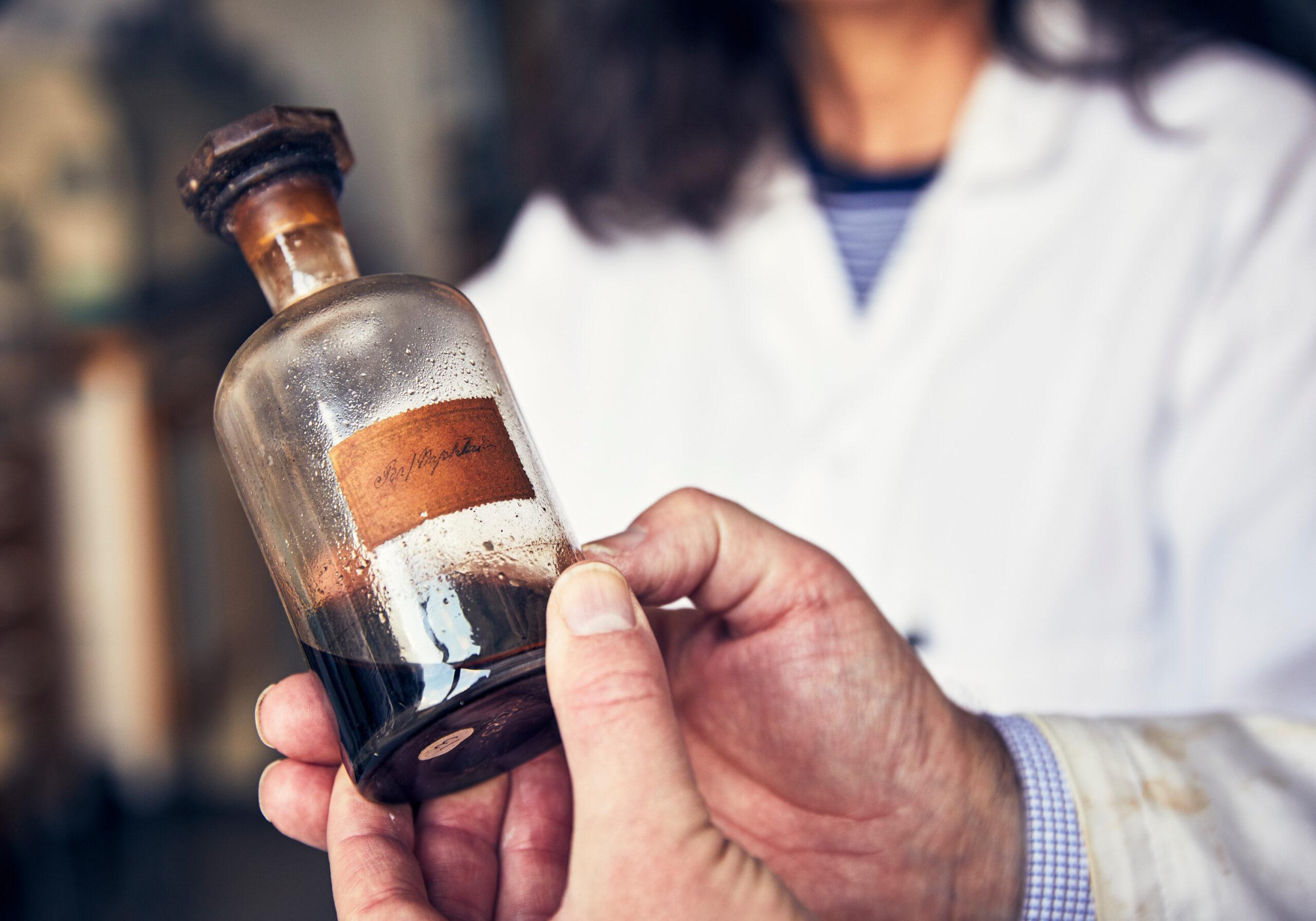 Two hands holding a glass bottle with an old label and some dark fluid inside.