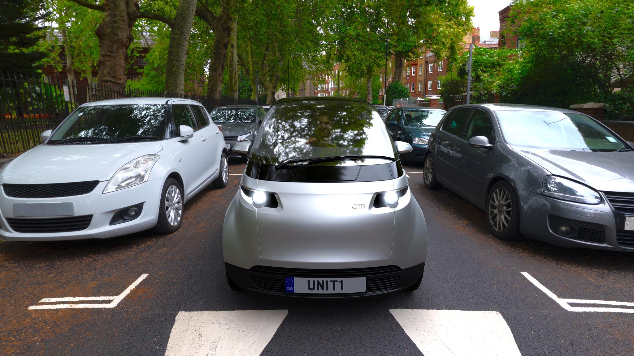A futuristic-looking, silver-coloured car driving on the road between parked cars on each side.