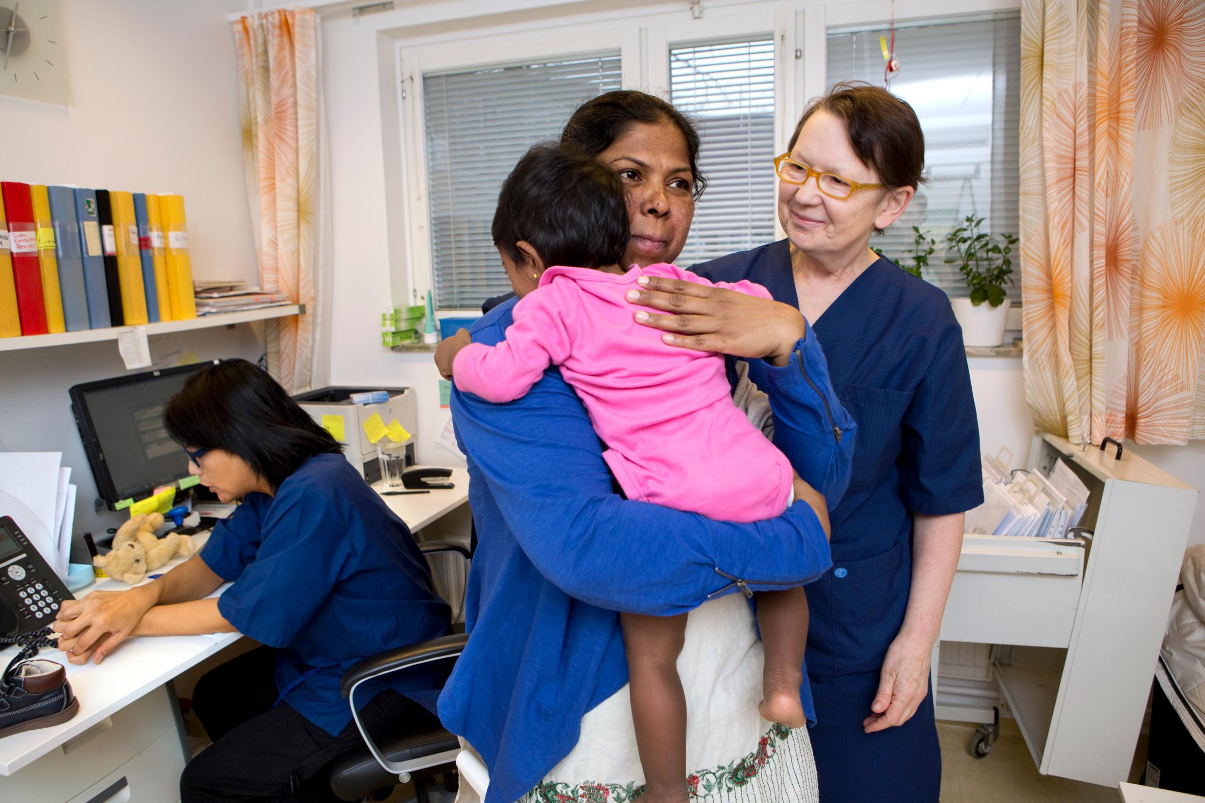 In the foreground is a mother holding a baby, slightly behind is a nurse and in the background is another person sitting at a desk, back towards the camera.