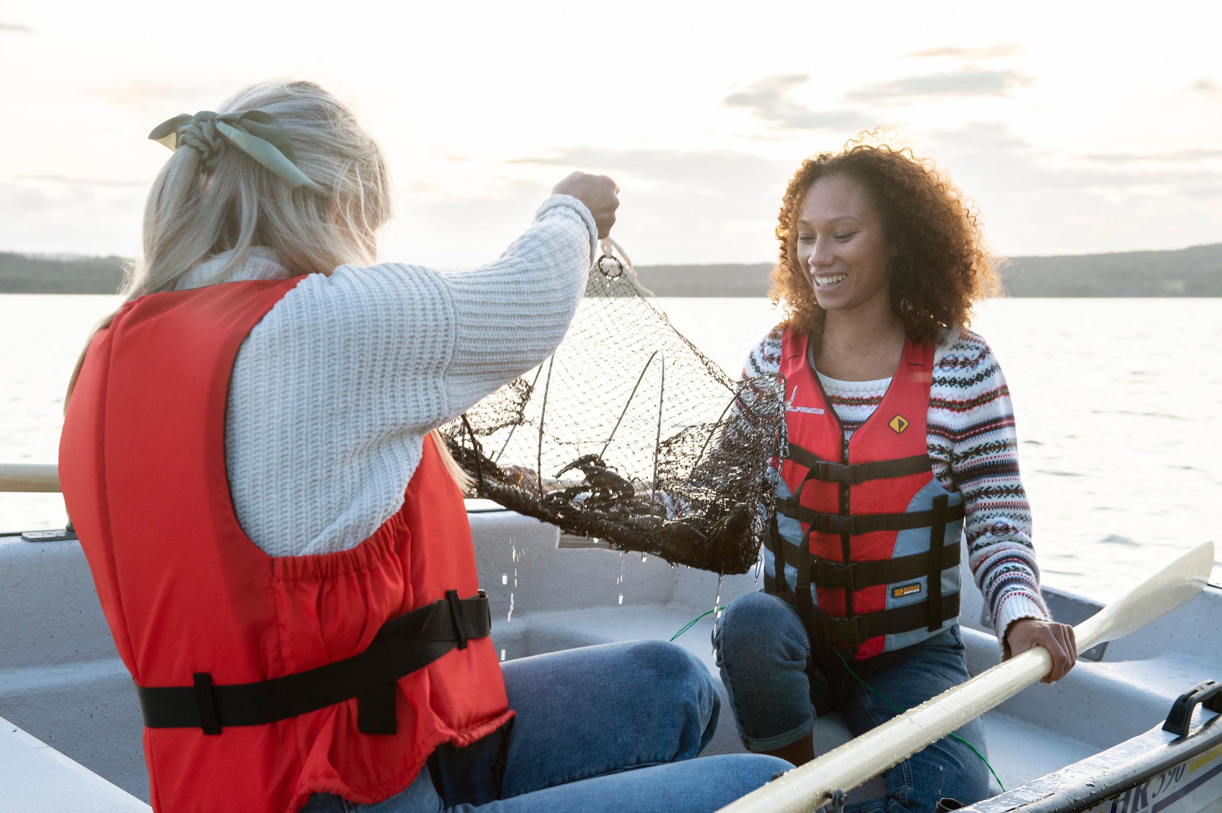 Two young women fishing crayfish from a small boat in a lake. They are holding a trap with some crayfish.