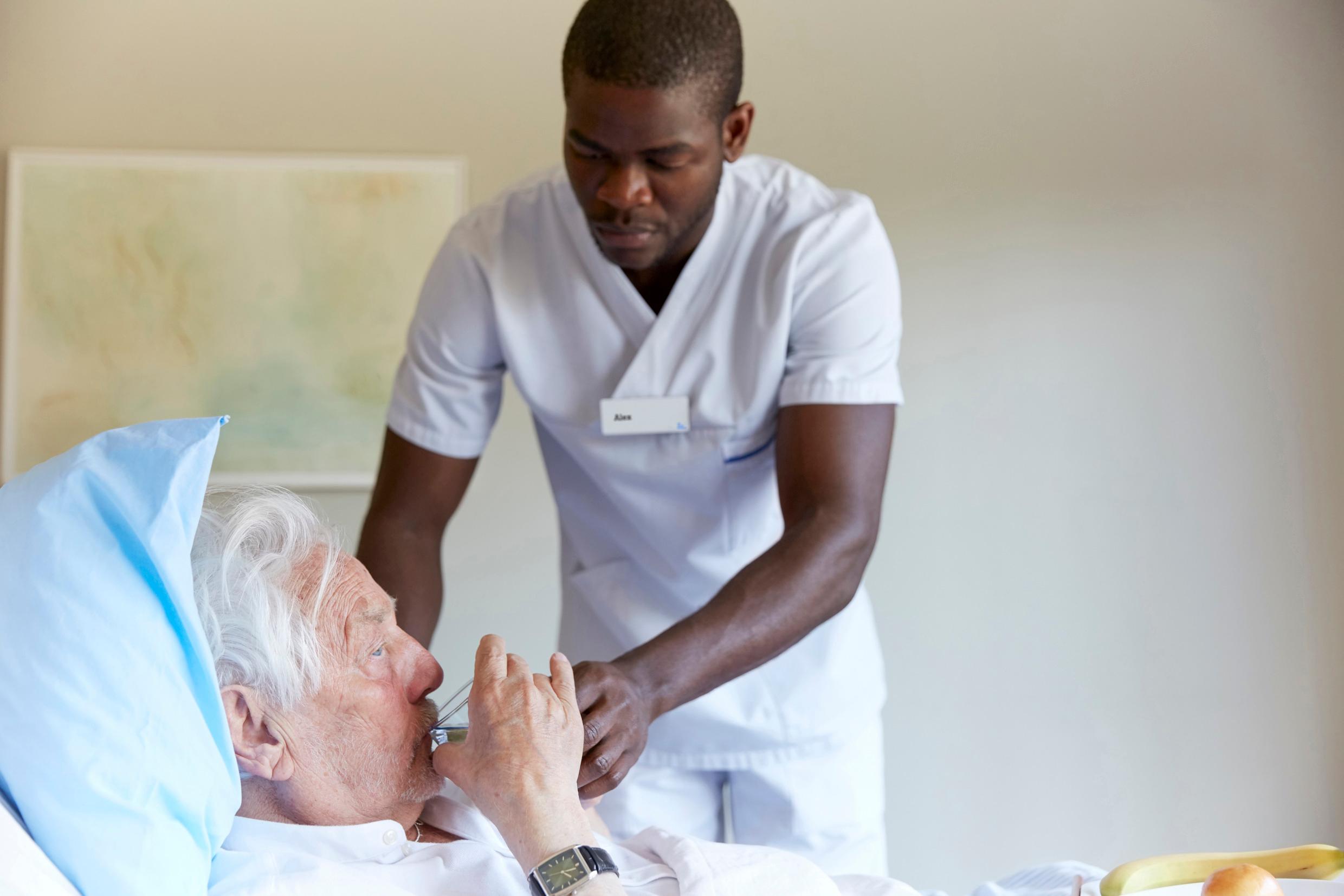 A male nurse in white scrubs tending to an elderly man half-lying in a hospital bed. An example of elderly care in Sweden.