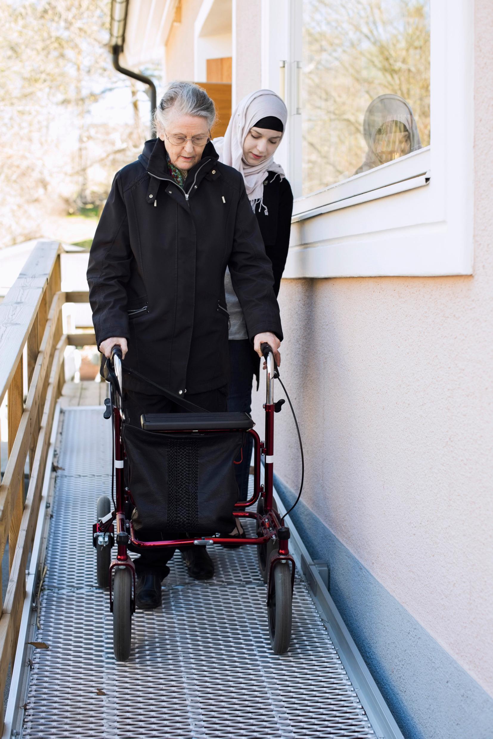 An elderly woman with a walking frame with wheels, being assisted by a younger woman who is behind her.