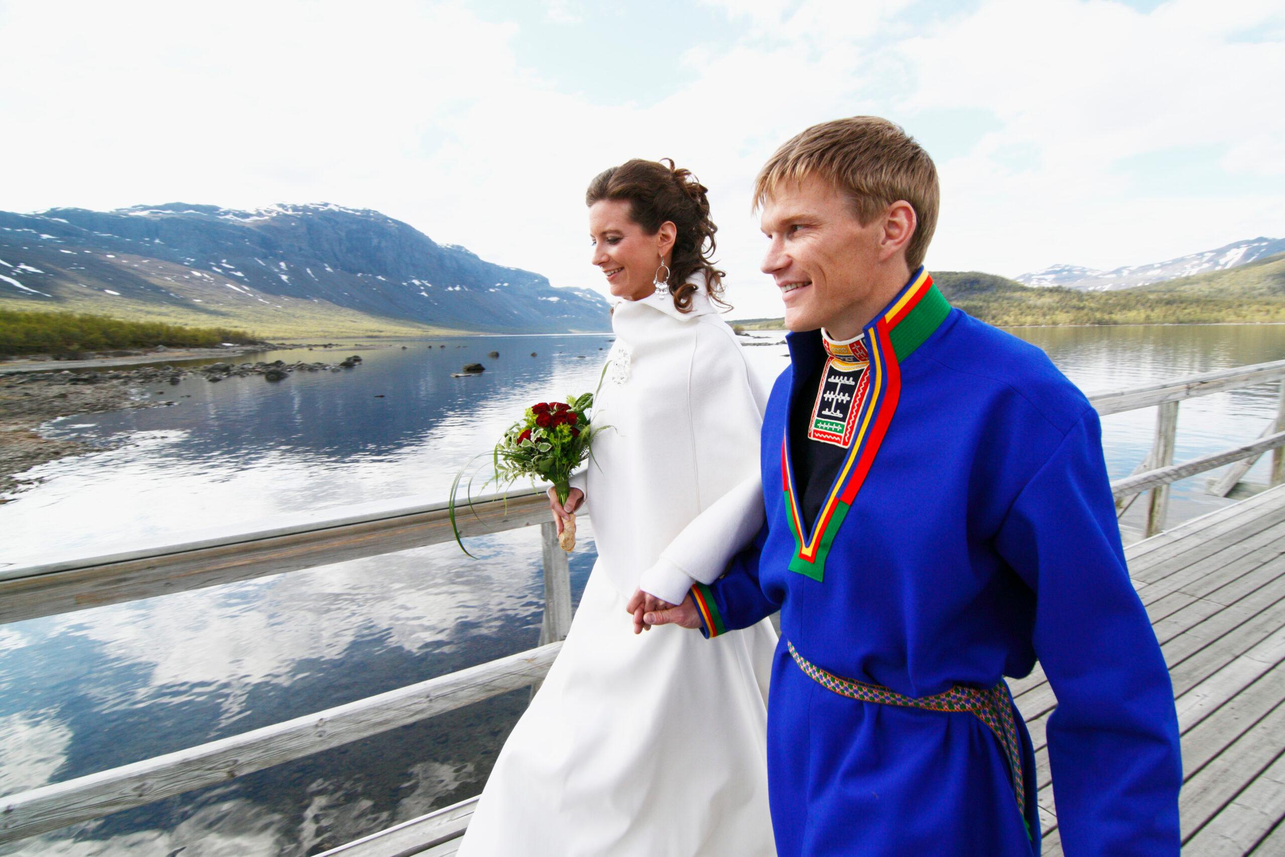 A newlywed couple - the woman in a white dress, the man in a traditional sami costume - walking across a bridge. Mountains in the background.