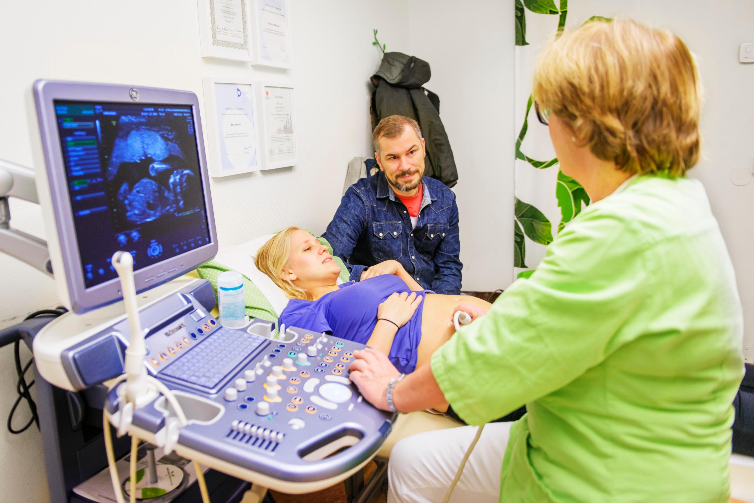 A woman lying down on an examination table, a man sitting behind her, and a medical person examining the woman, her back to the camera. On the left is a computer screen.