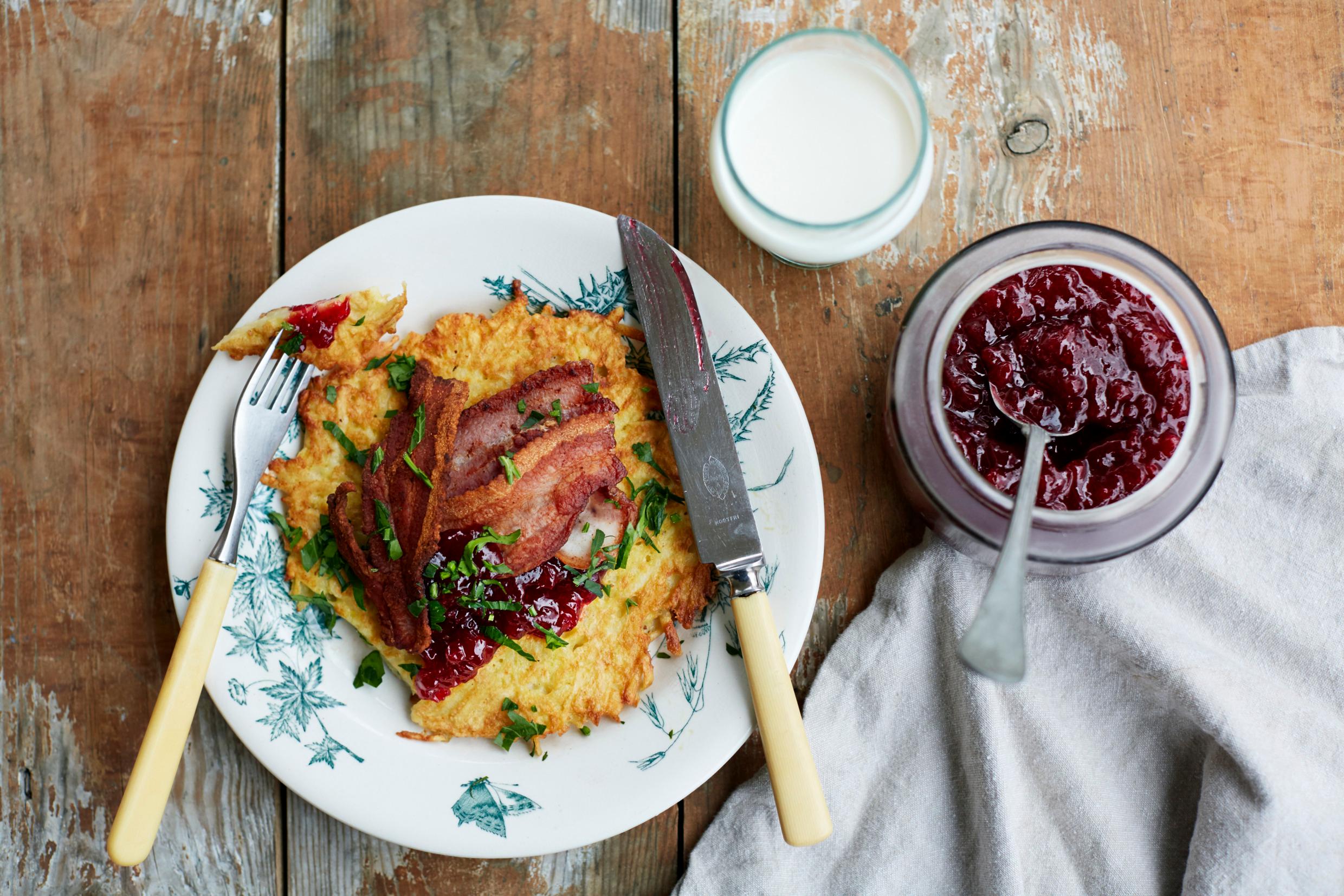 Seen from above, a wooden table is set with a plate of potato pancakes and bacon, a jar of lingonberry jam and a glass of milk.