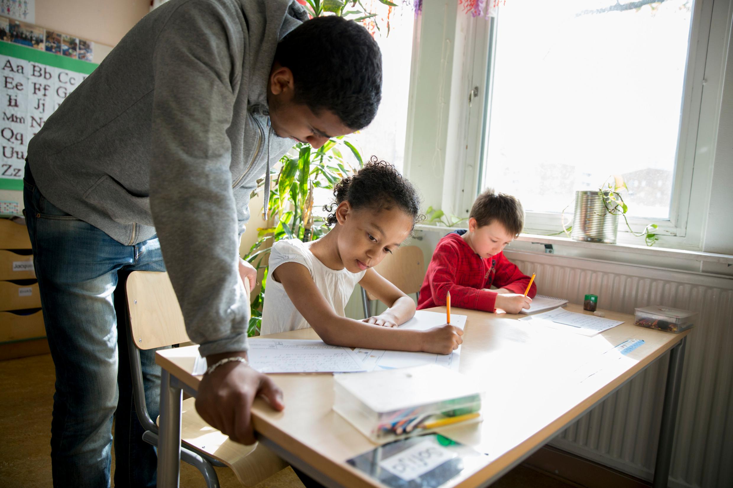 A classroom with two children sitting at their desks, a male teacher leaning over one of them.