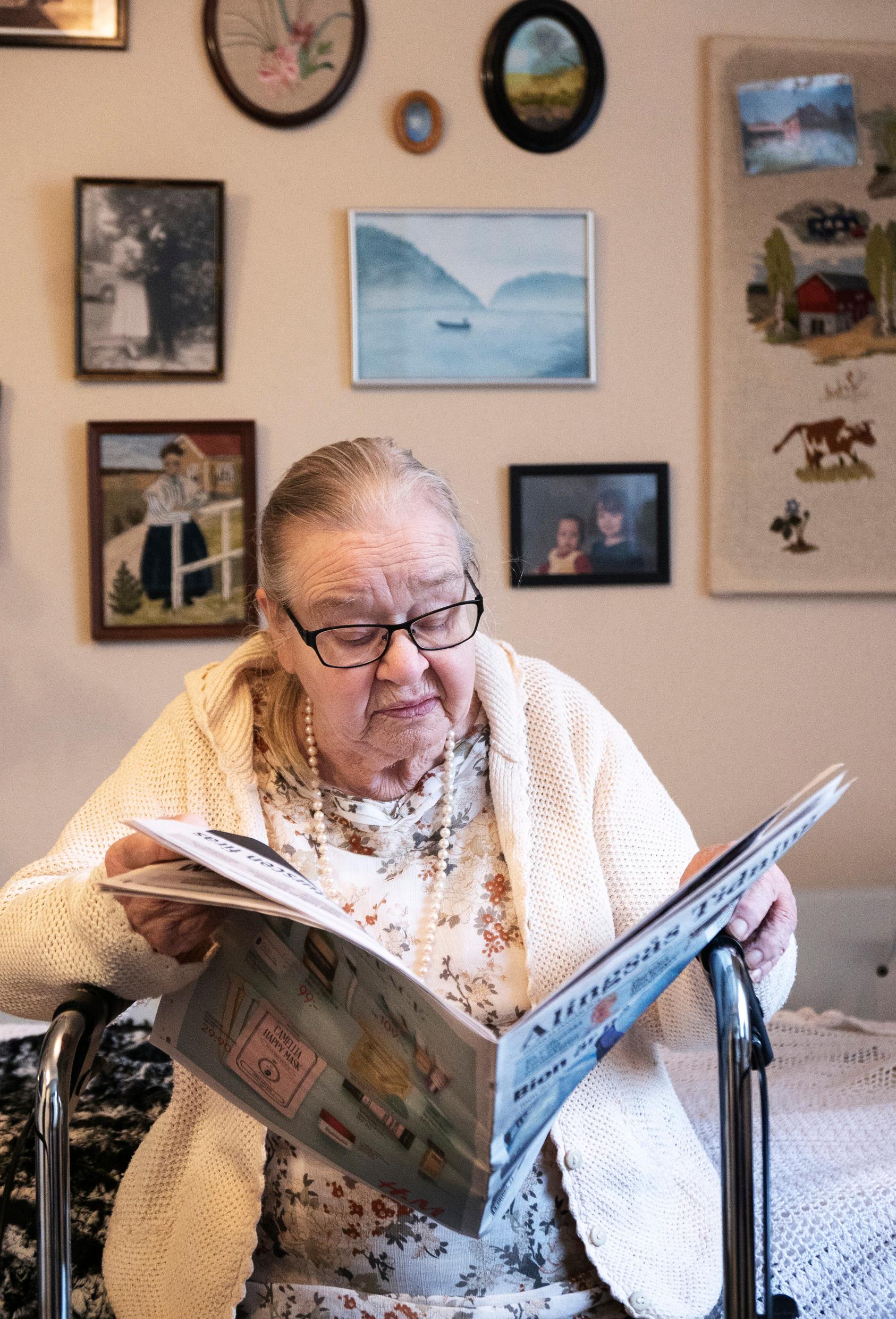 An elderly woman sits on a bed, leaning on a walker, reading the paper.