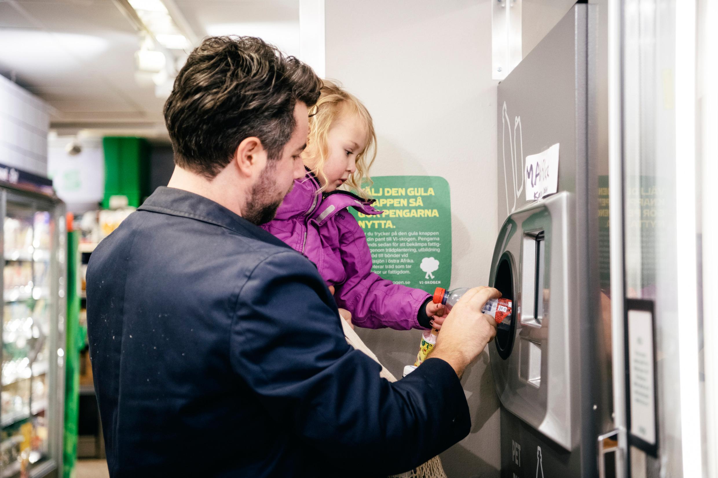 A man is holding a small child. Together they push a plastic bottle into a machine,