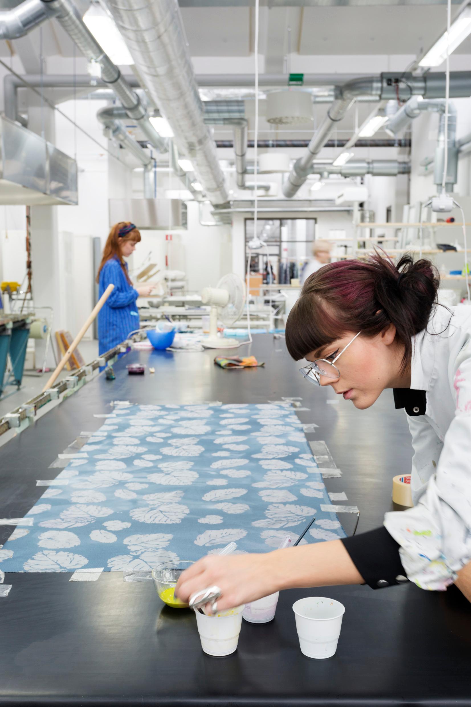 A woman leaning over a piece of leaf-patterned fabric, another woman in the background.