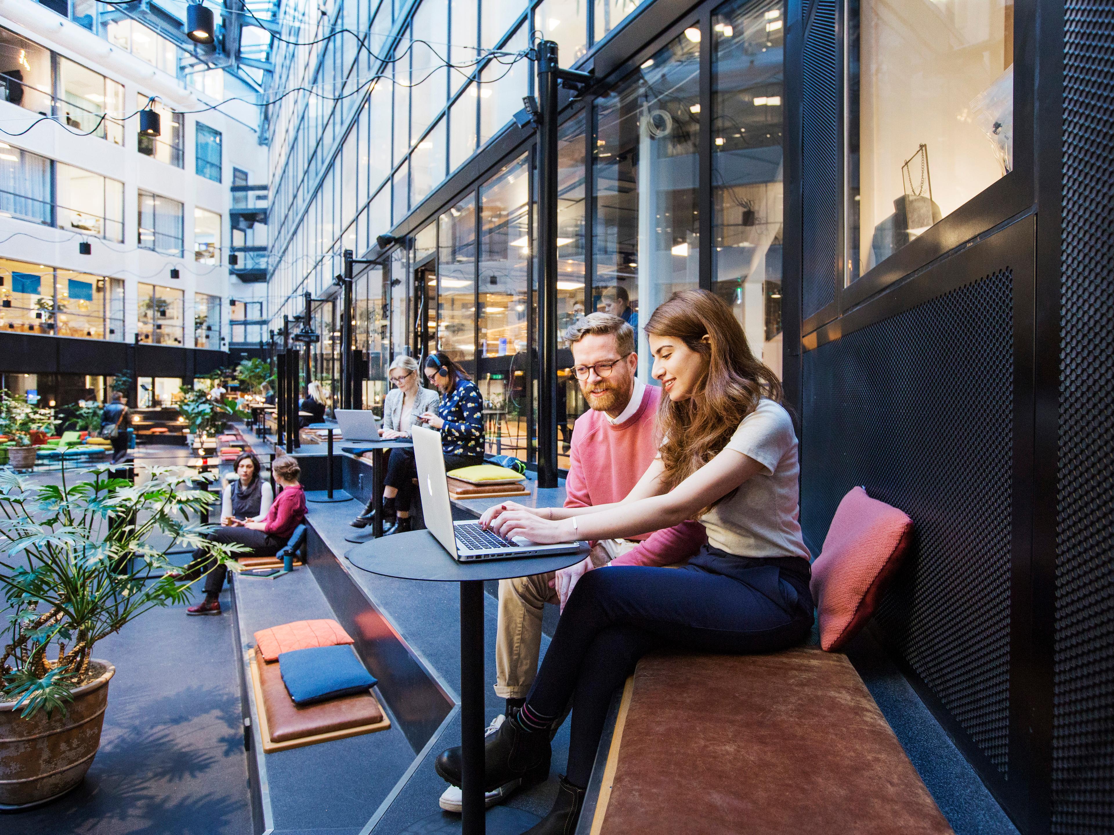 A man and a woman seen from the side. They sit on a bench looking at a computer screen. Four other people in the background.