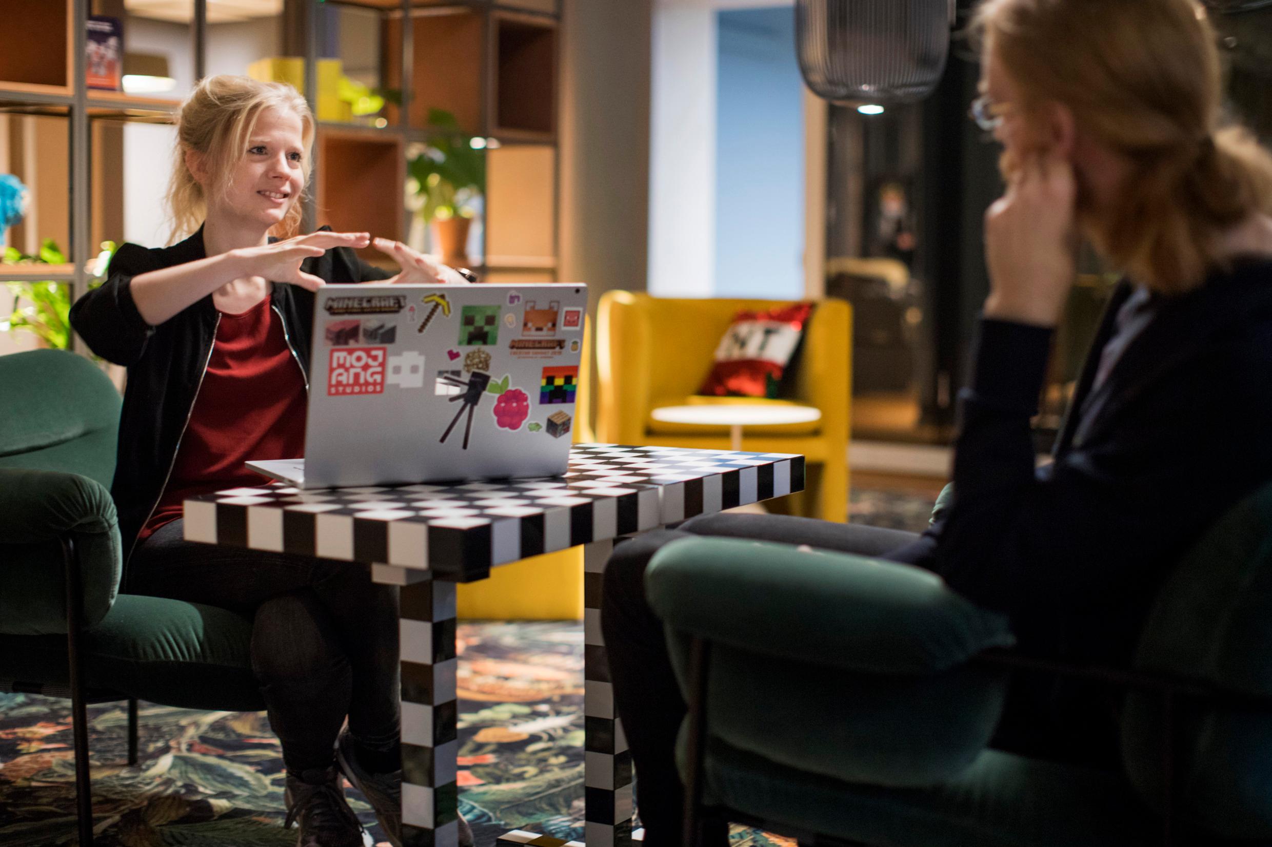 A man and woman sit in a creative offfice environment. There's a laptop on a table between them. The woman gestures something with her hands.