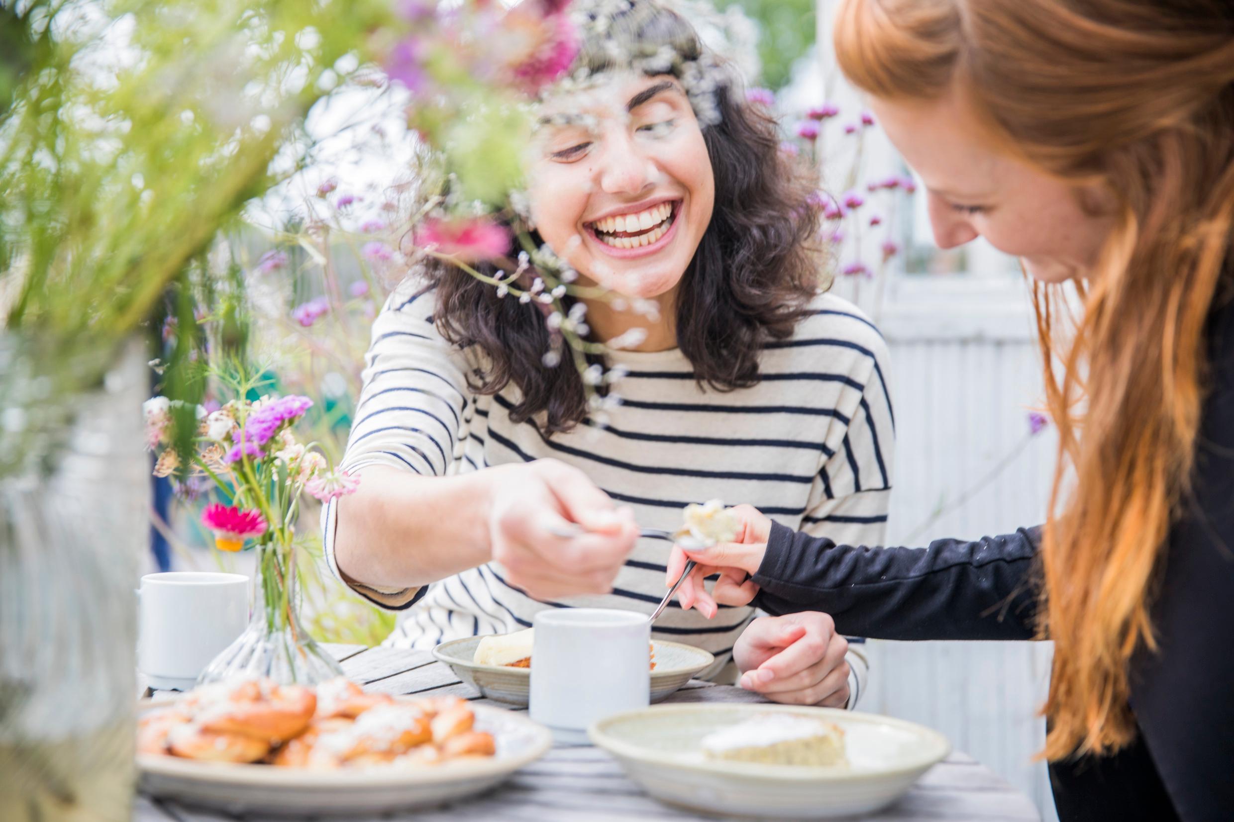 Two women having an outdoor fika. They drinking coffee and sharing cake with each other.