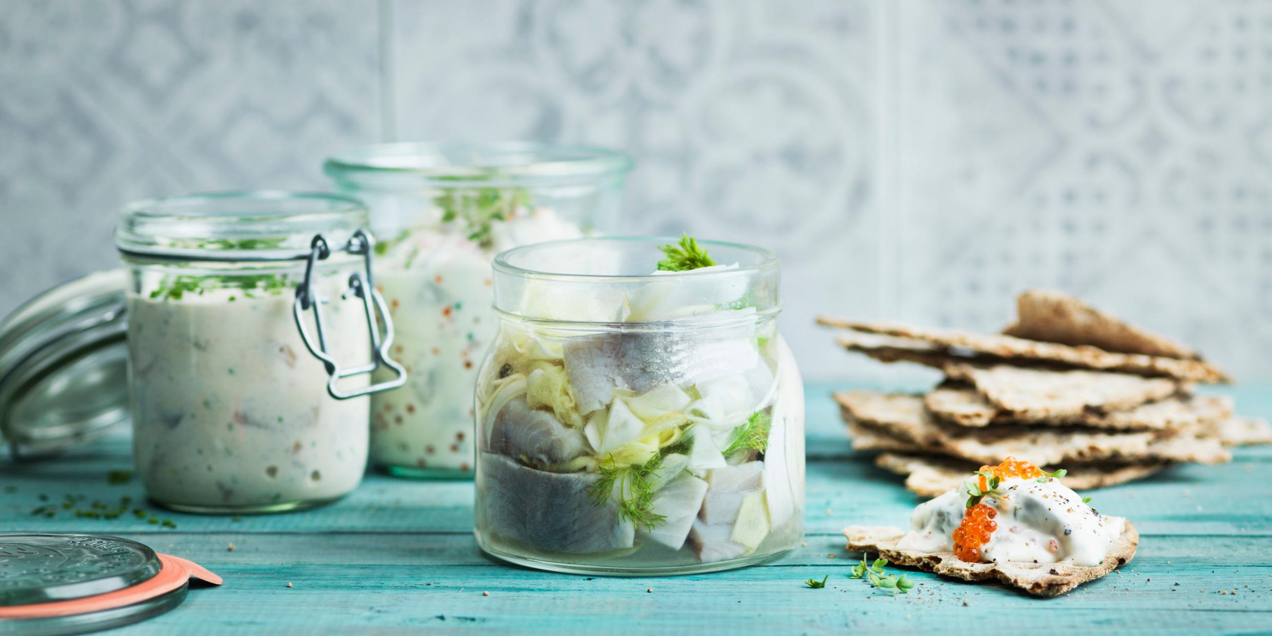 Three jars with pickled herring on a wooden table next to some crispbread an a piece of crispbread with some pickled herring topped with fish roe.