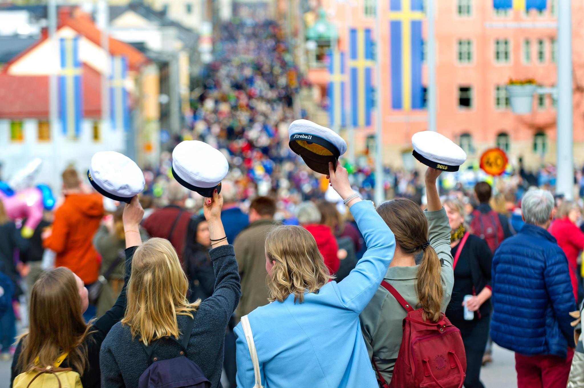 Large crowds of people in a city. In the foreground are four girls with their right arm lifted, holding a white cap.