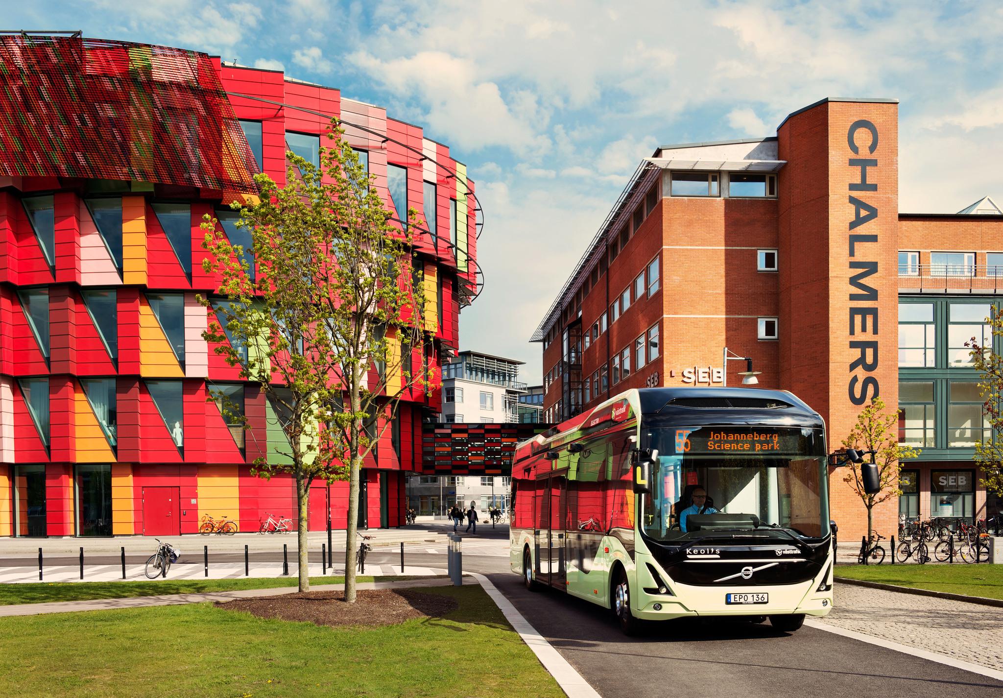 A bus driving on a street with houses in the background. To the left, a colourful rounded building and to the right, a brick building with a sign reading 'Chalmers'.