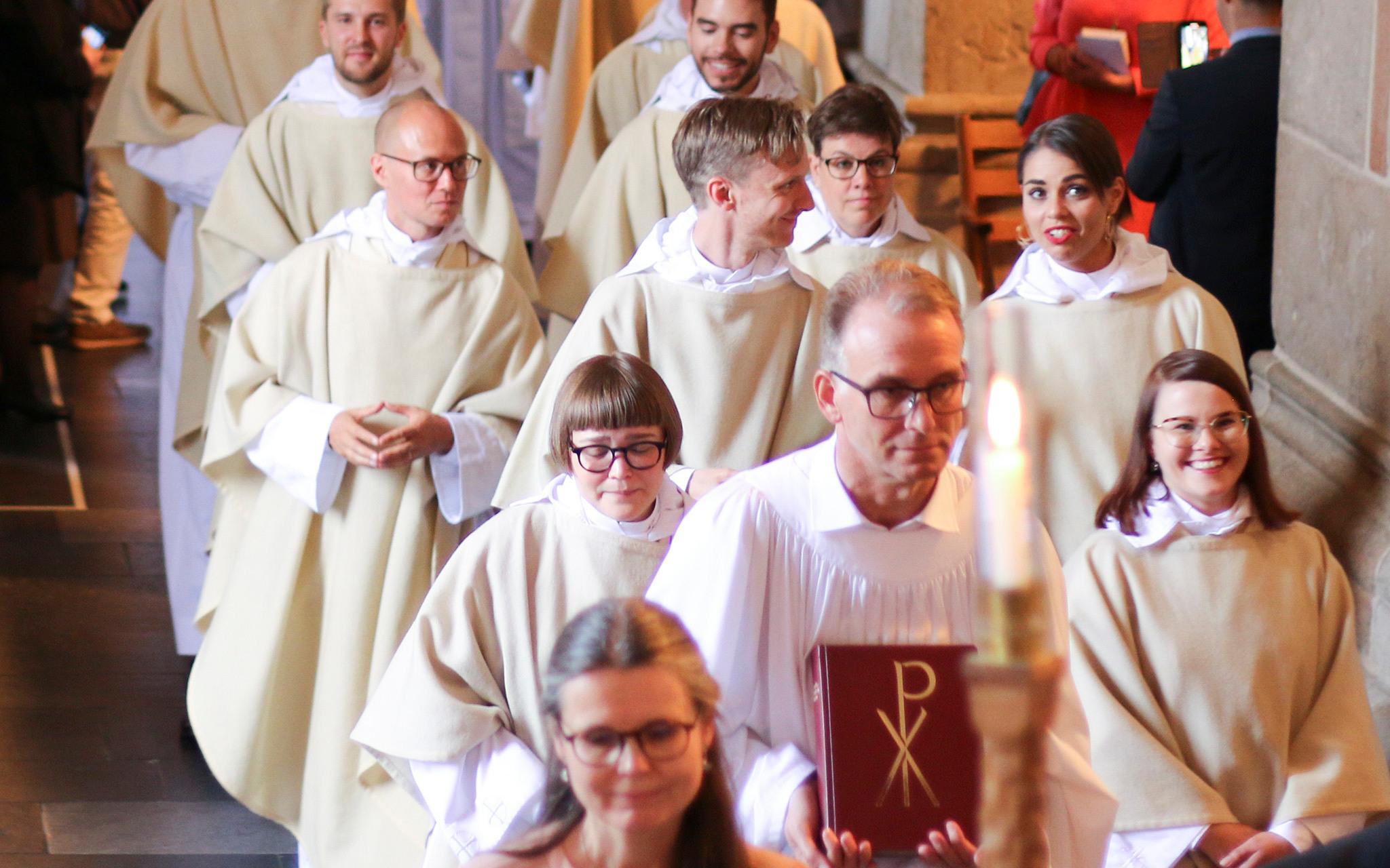 Priests to be walking through a church dressed in long white cloaks. At the front is a man carrying a bible.