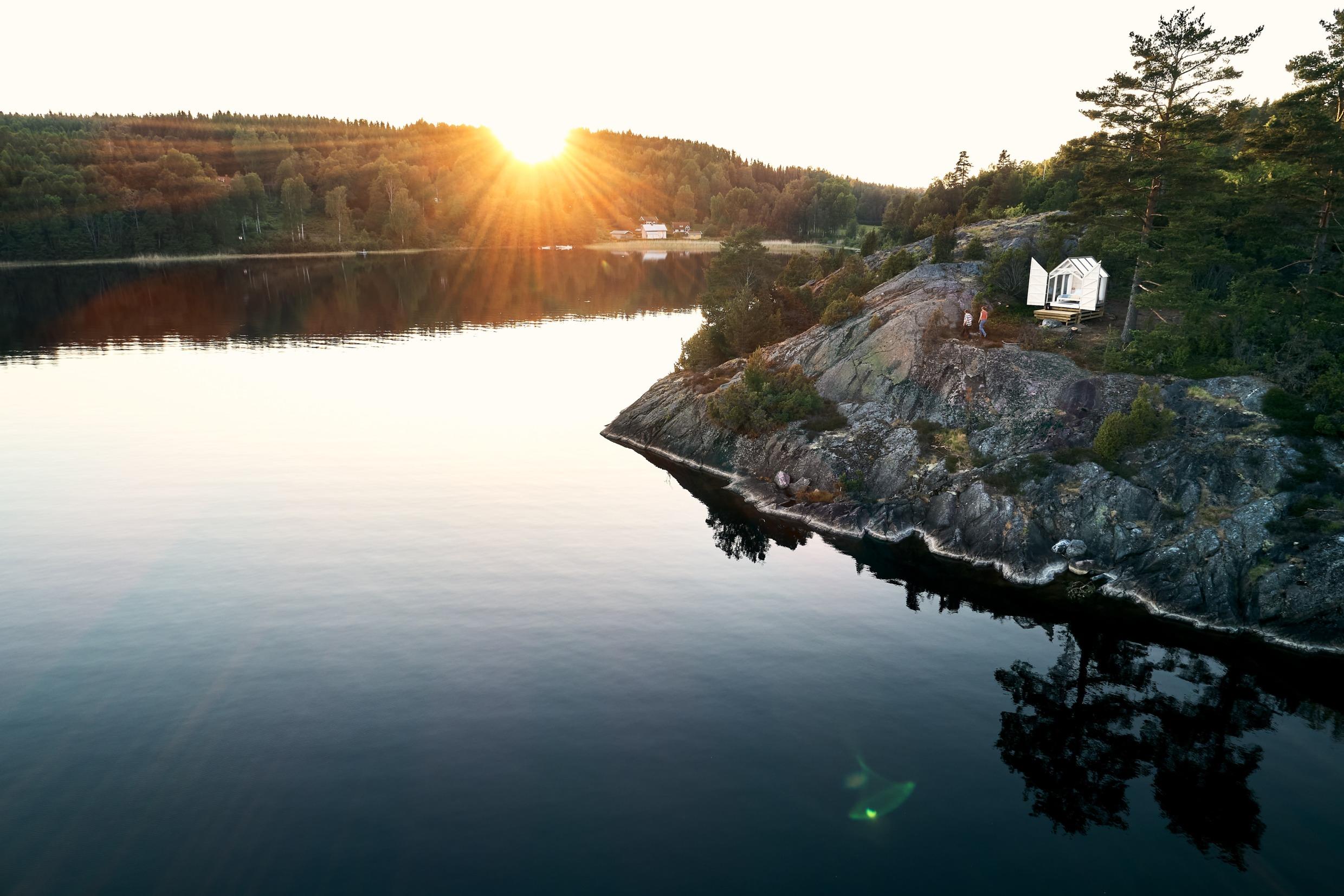 View of a glass cabin on a cliff by a lake. Two people are walking in front of the cabin. The sun is setting behind the forest.