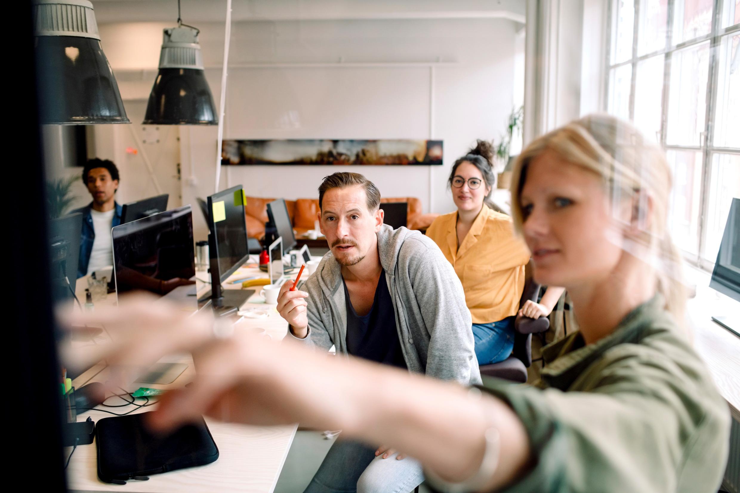 People around a desk are looking at a screen. A woman is pointing at the screen (out of sight).