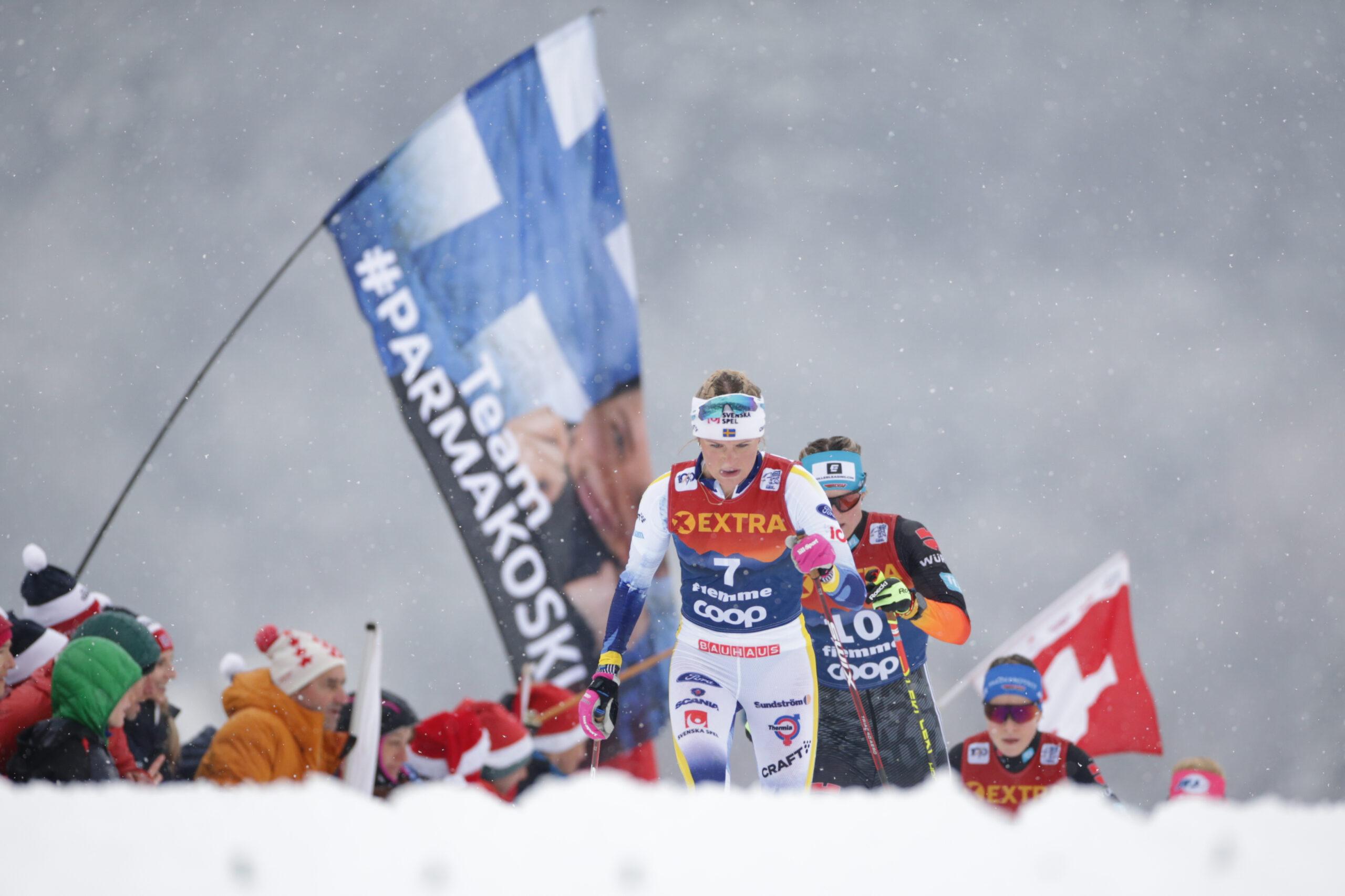 Skiers on snow, a Finnish flag in the foreground.