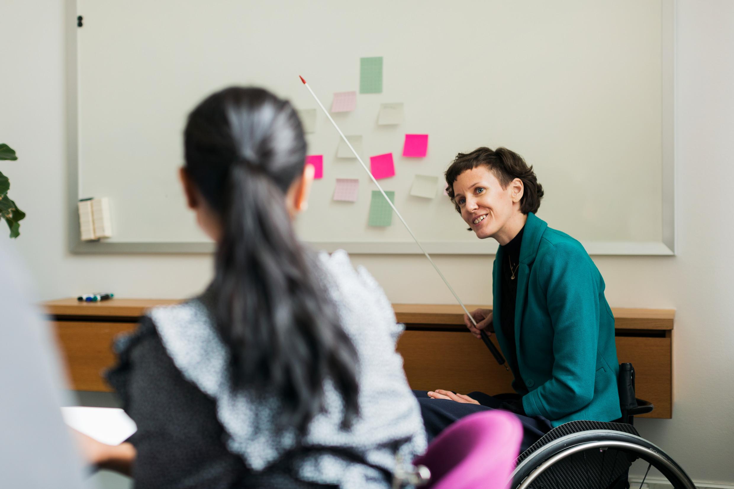 A woman in a wheelchair is pointing at a whiteboard in an office environment.