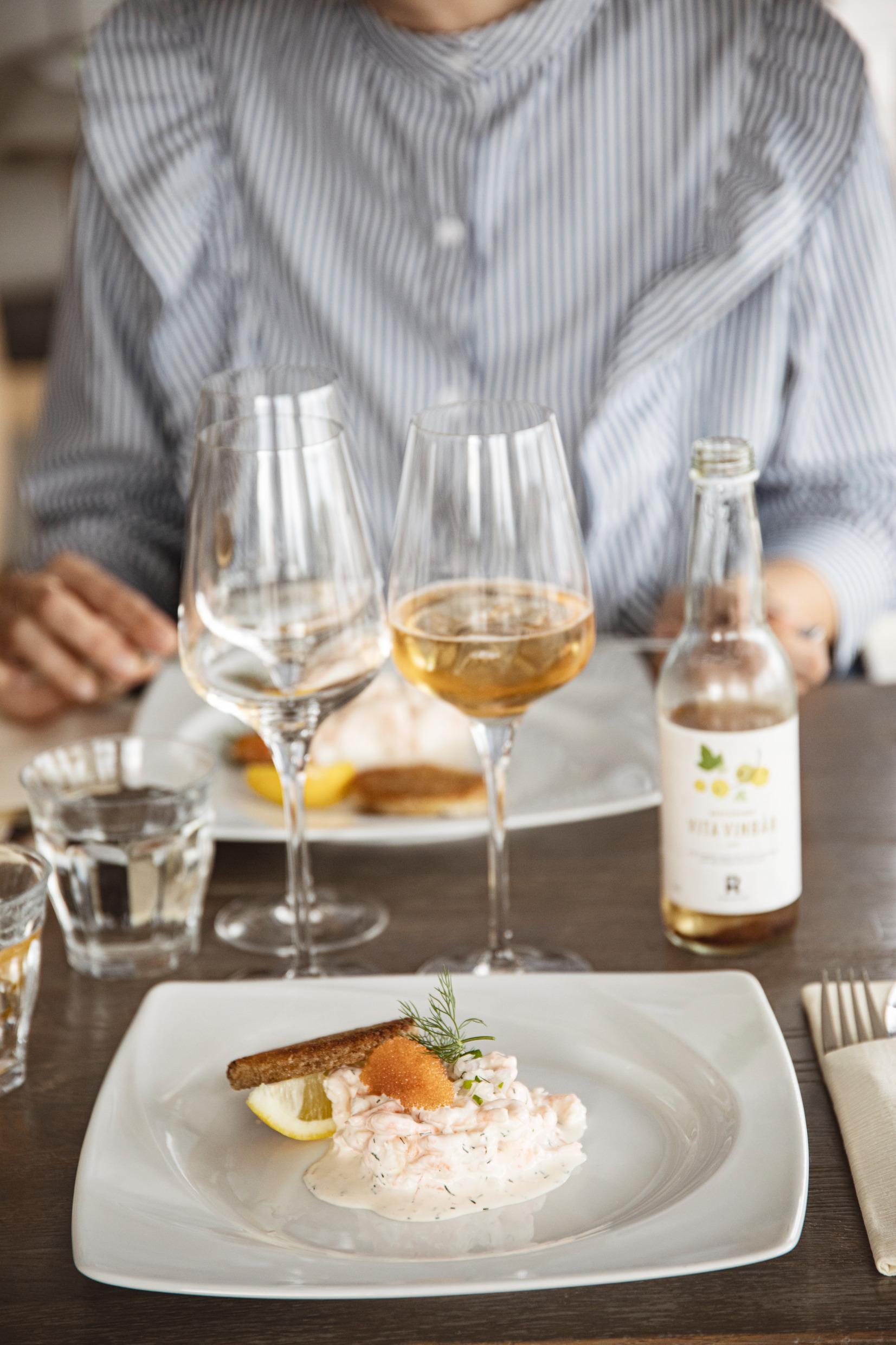 A white plate with Skagen with roe and dill. Next to it is a piece of bread and a lemon slice. A person sits at the other side of the table.