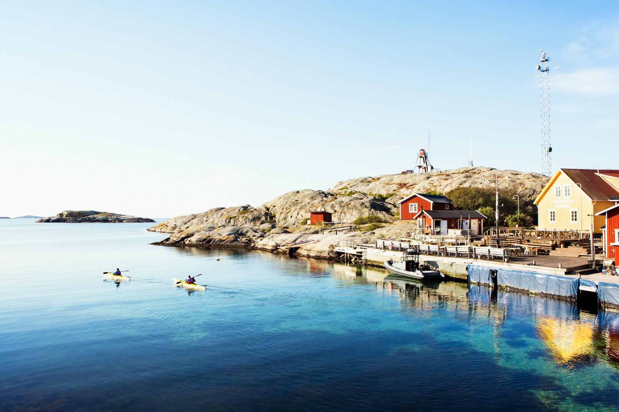 An island with some small houses. In front of it, two people in kayaks in the sea.