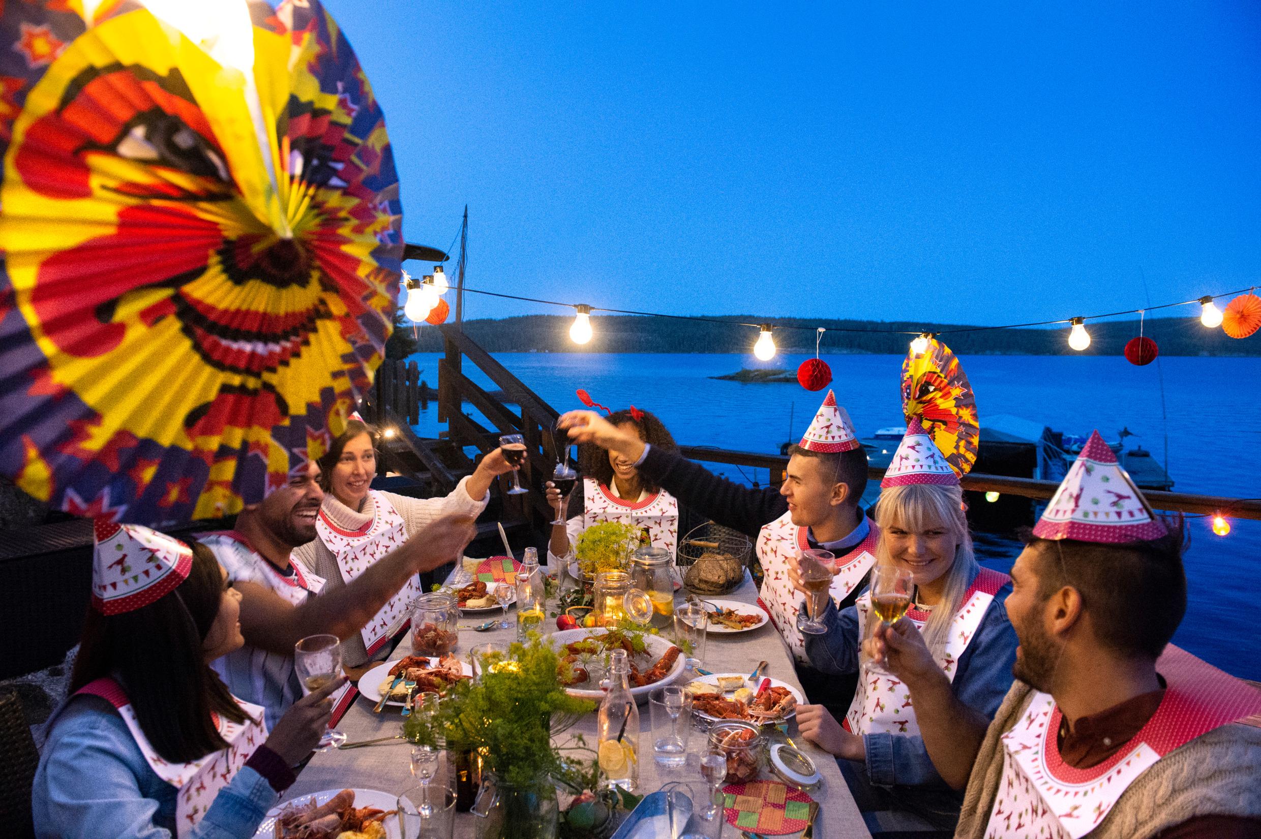 Crayfish party at a jetty by the sea. Seven peoples around a table dressed in paper hats and bibs are eating crayfish and toasting with beer and wine. There are paper lanterns hanging above the table.