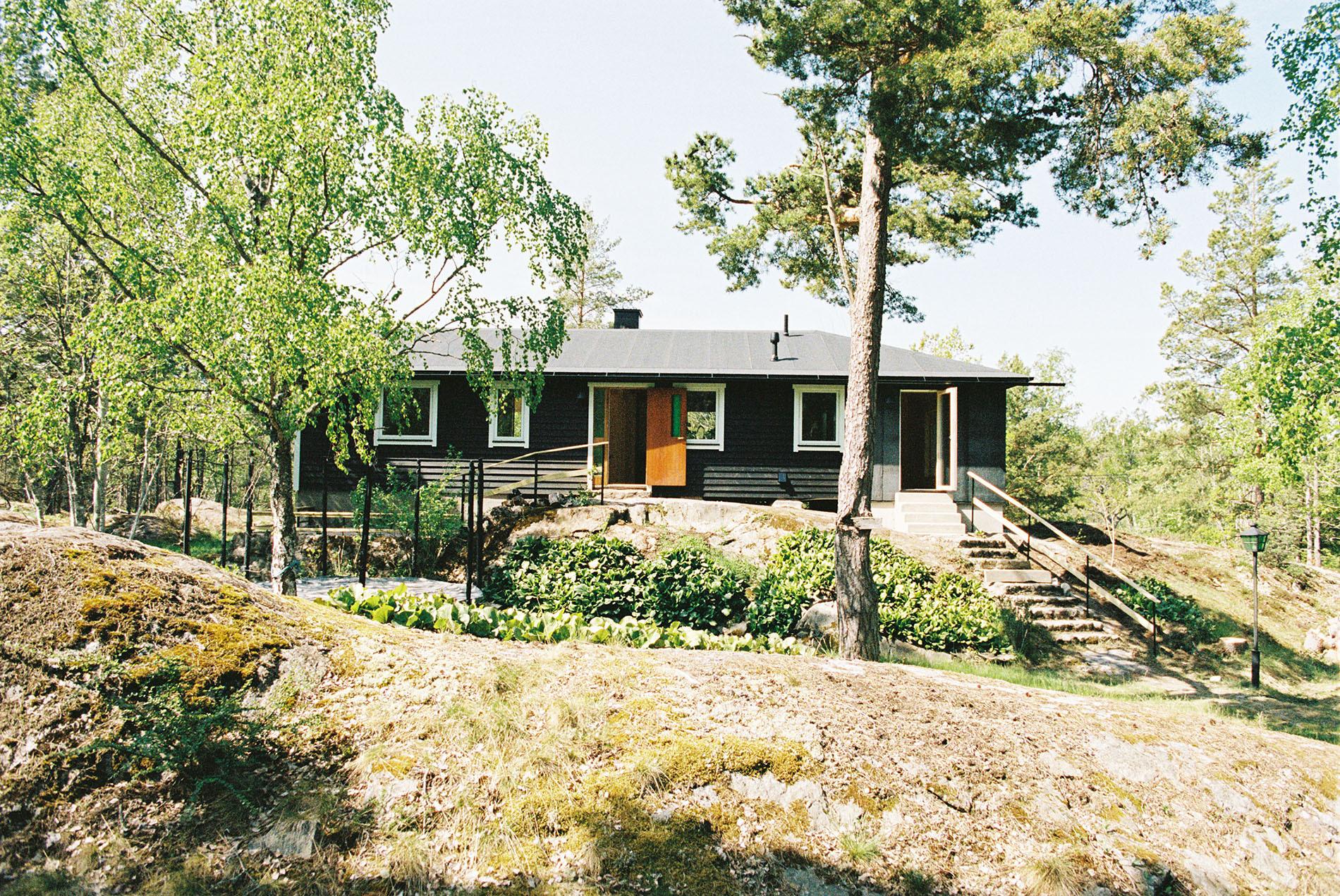 A low, dark-brown house on a rocky hill among some trees.