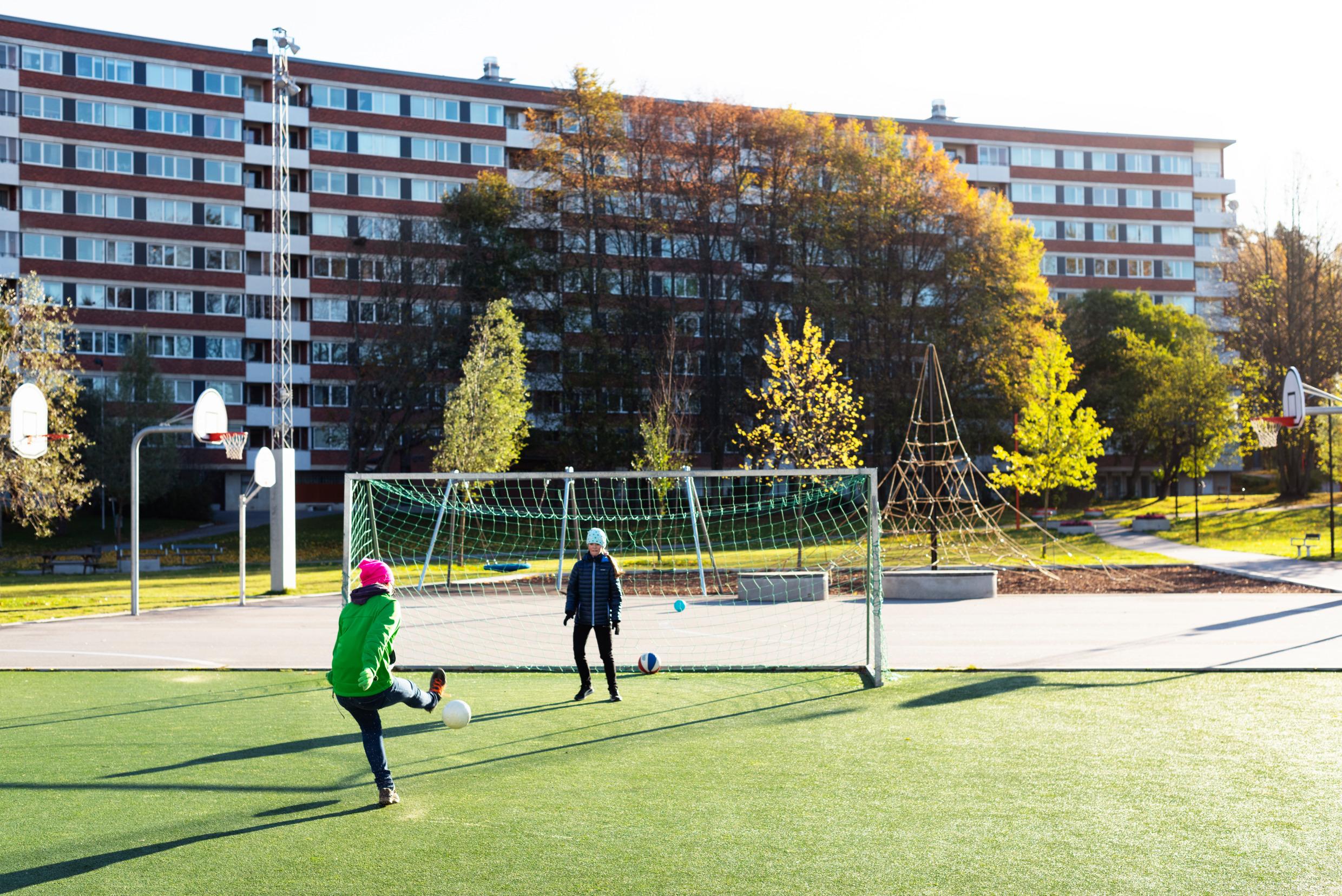 Two girls are playing football outside some apartment buildings in an area with a football pitch, basketball courts and a playground.