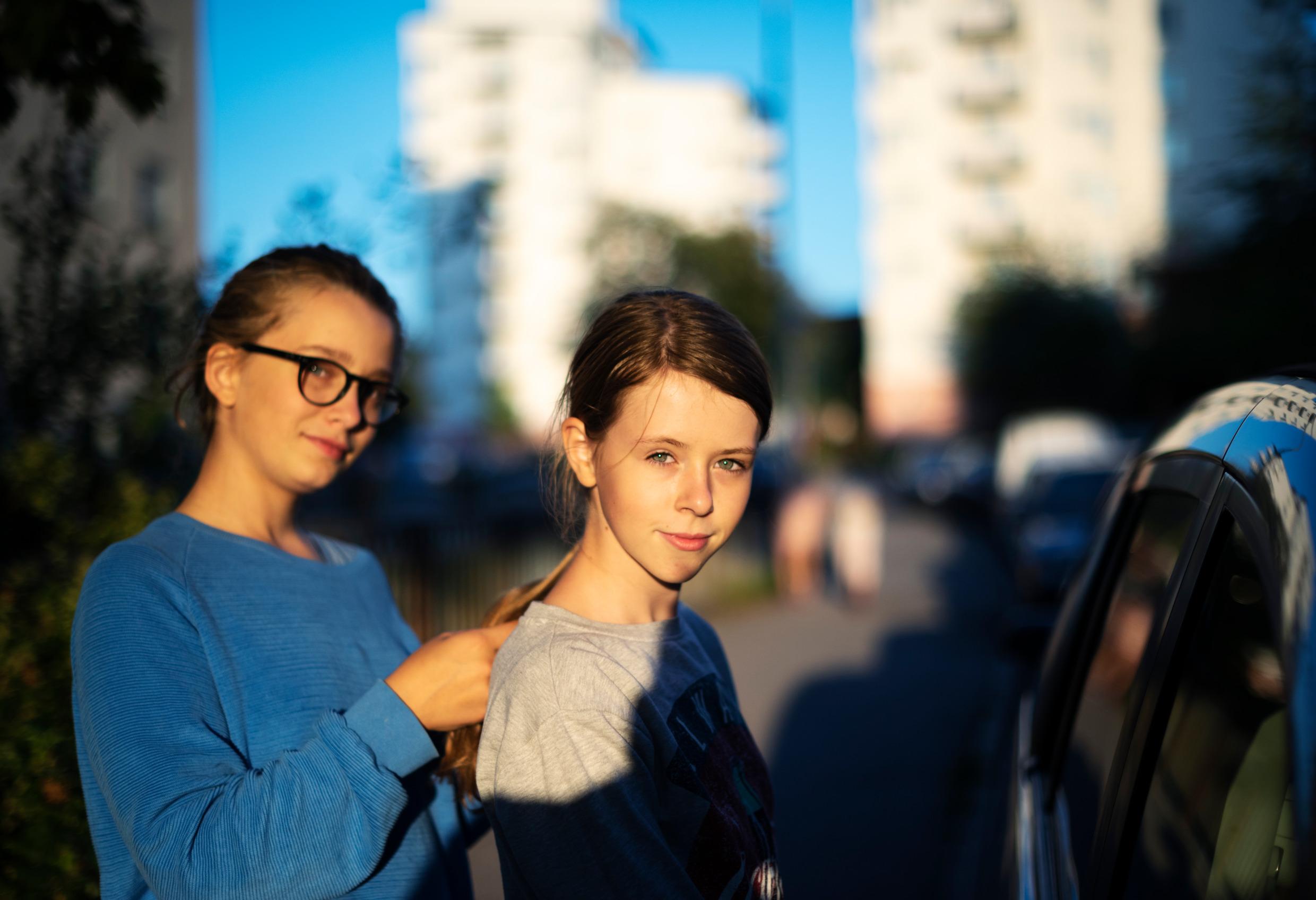 A girl is braiding her friend's hair. There are apartment buildings in the background.