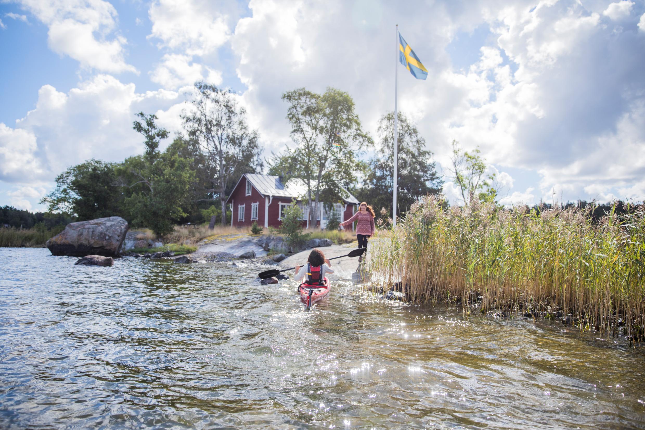 A woman on a cliff receives another woman who comes paddling in a kayak in the archipelago. In the background a red cottage and a Swedish flag.