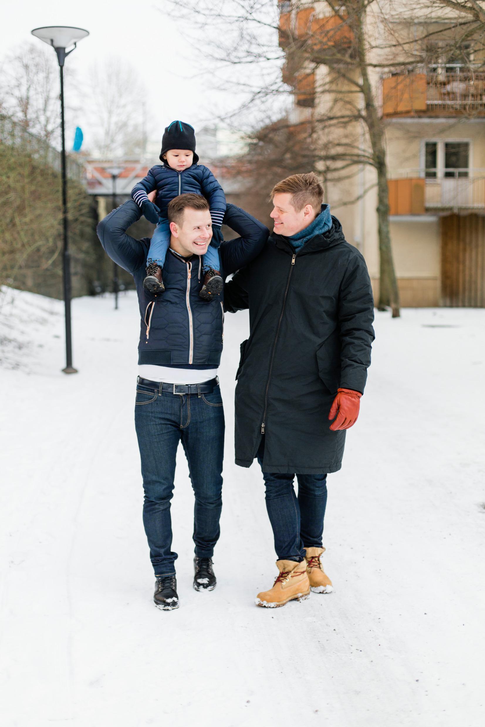 Two men walk down a snow-filled street, embracing, one lifting a child over his head.