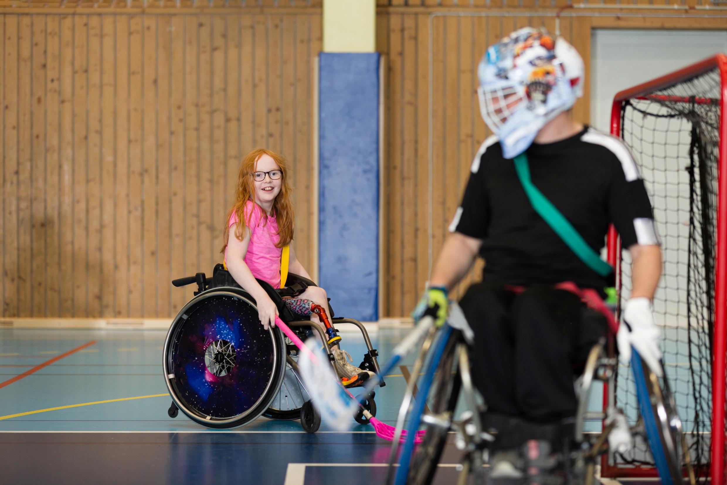 A girl plays wheelchair floorball, in the foreground a male goalkeeper in a wheelchair.