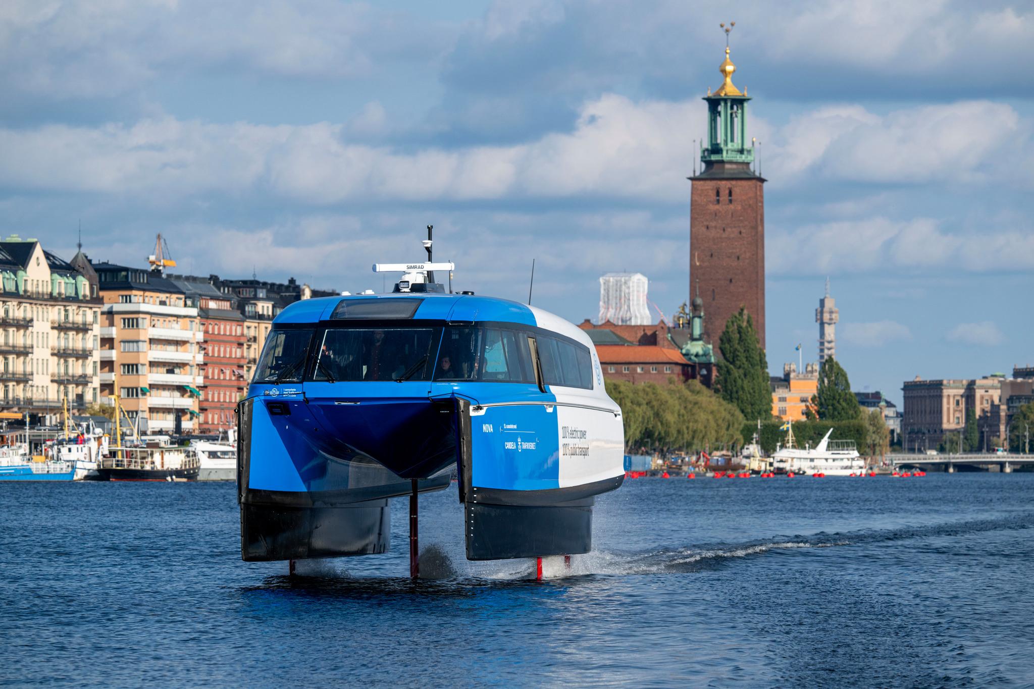 A blue and white boat seemingly flying over the surface of the water. Stockholm City Hall in the background.