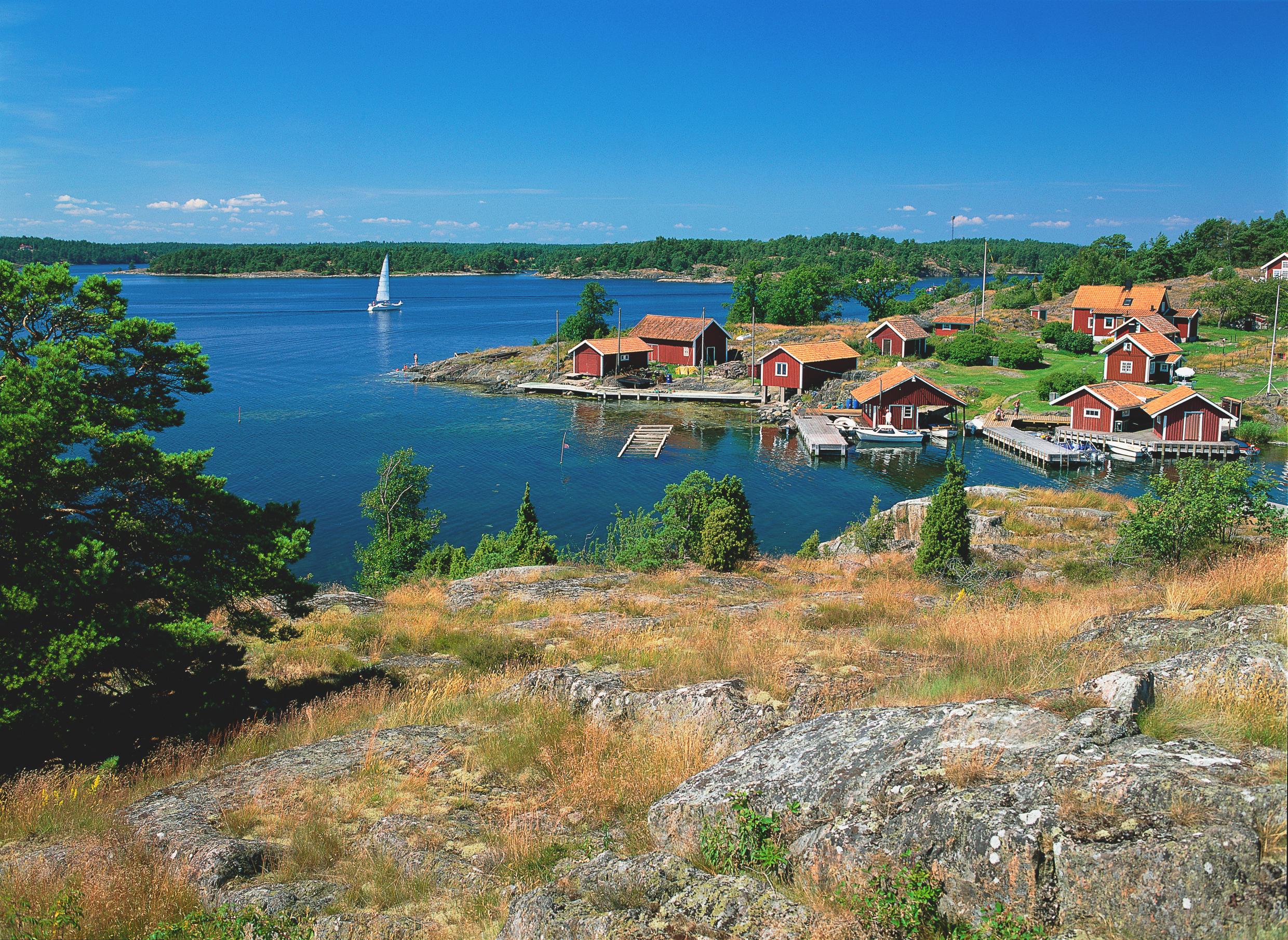 Red cottages on an island in the archipelago on a sunny day. A sailboat sails by the islands.