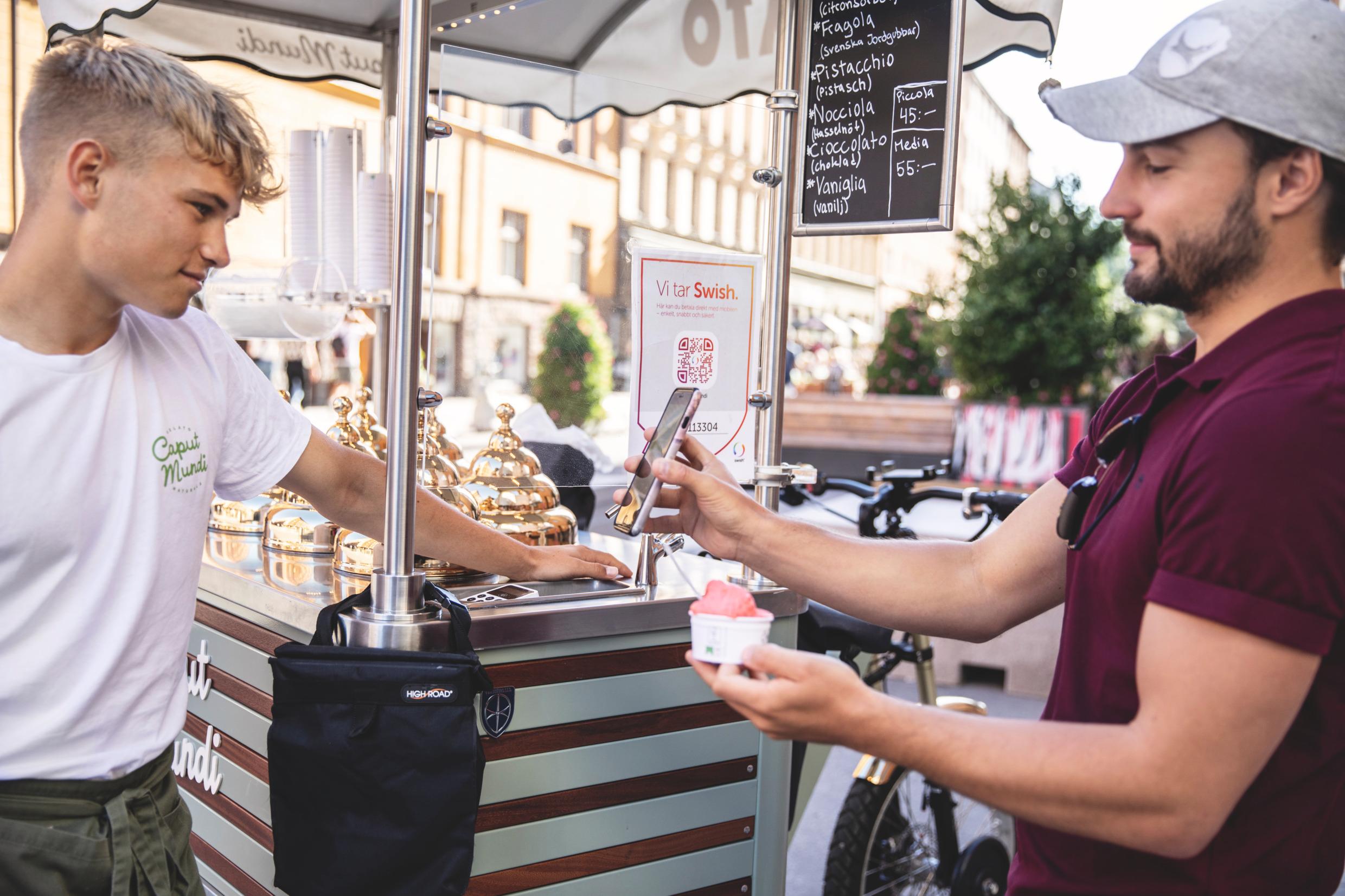 A young man in t-shirt and caps buys ice cream and pays with his smartphone.