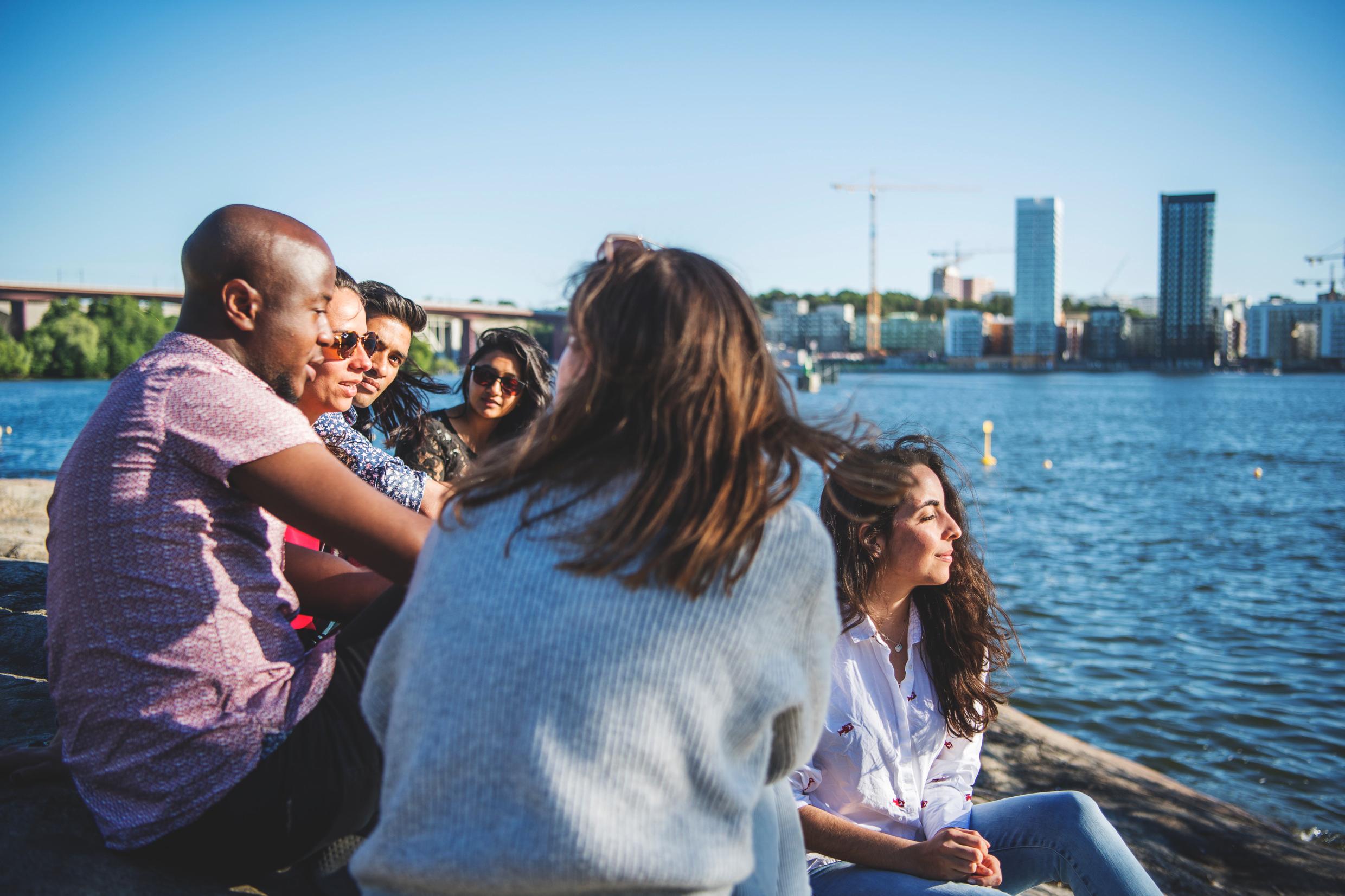 People sitting together by the water in Sweden, with a city skyline in the background.