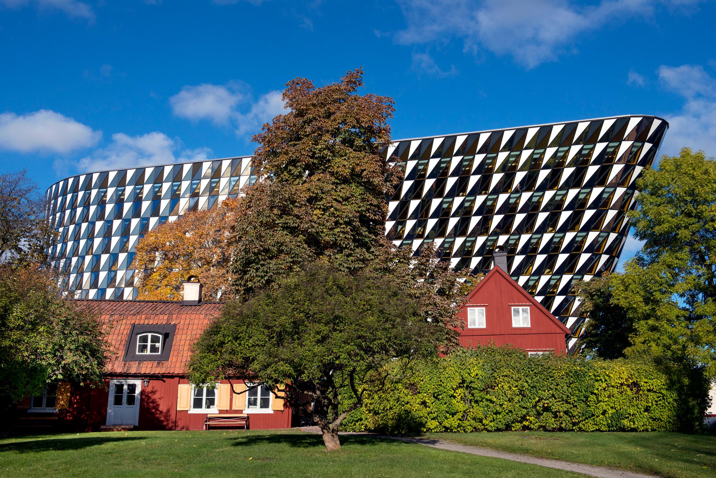 A supermodern geometrical building behind traditional Swedish red wooden houses in a park with green grass and trees, blue sky in the background.
