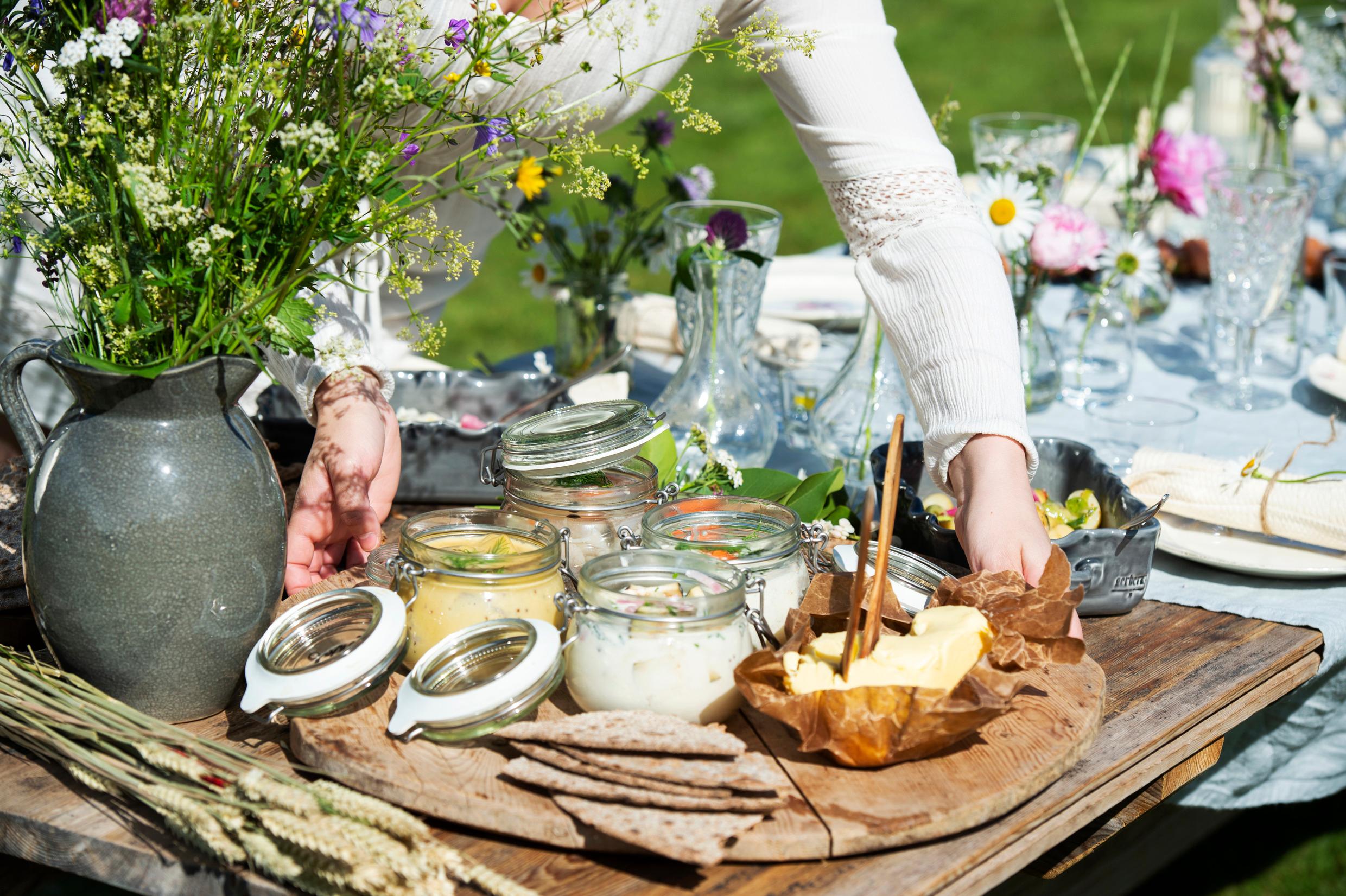 Herring in glass jars, butter and crispbread on tray on a table. Two arms are seen holding the tray, a vase with flowers on the left.