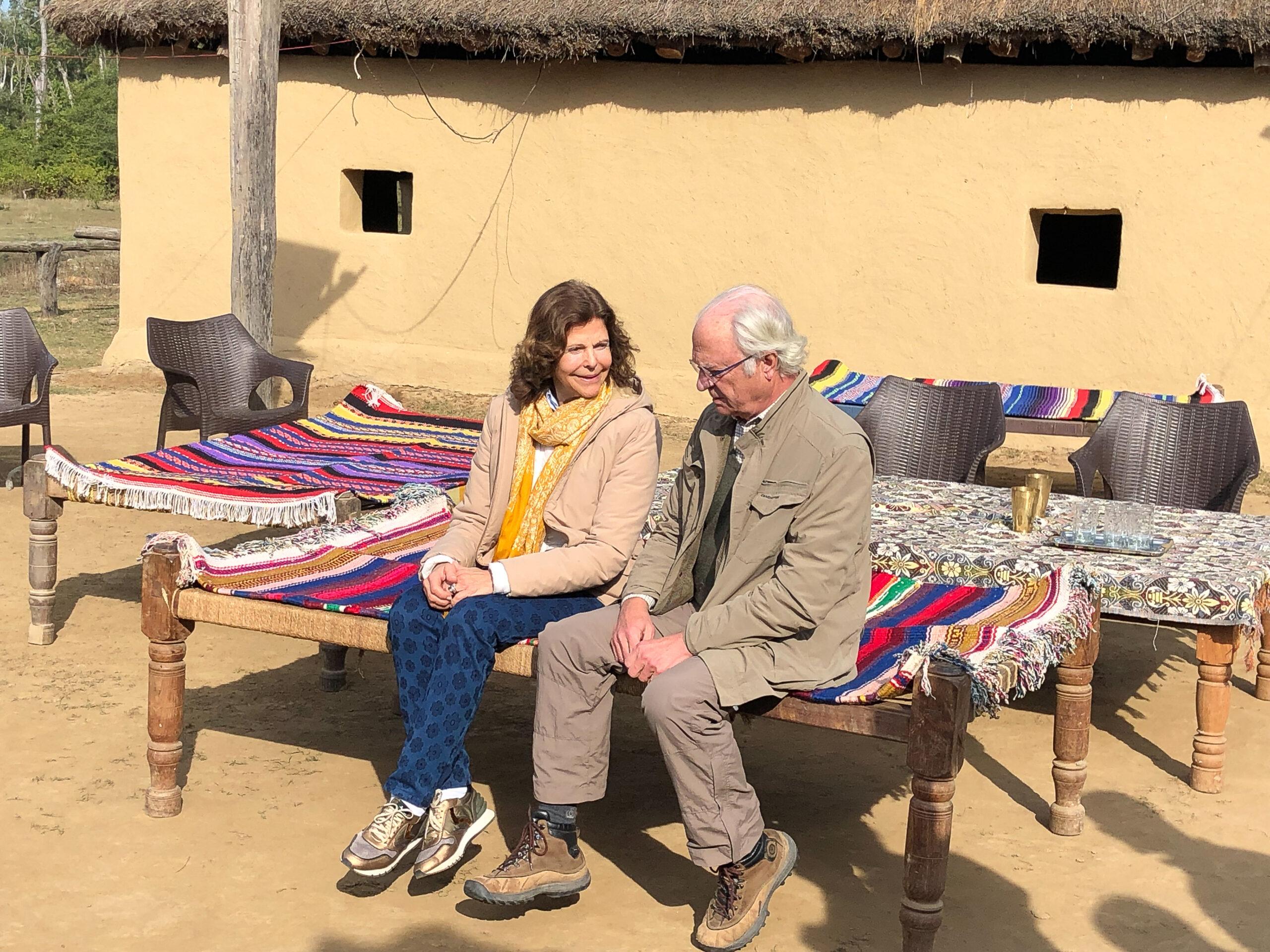 Queen Silvia and King Carl XVI Gustaf sitting on a bench in leisure clothes.